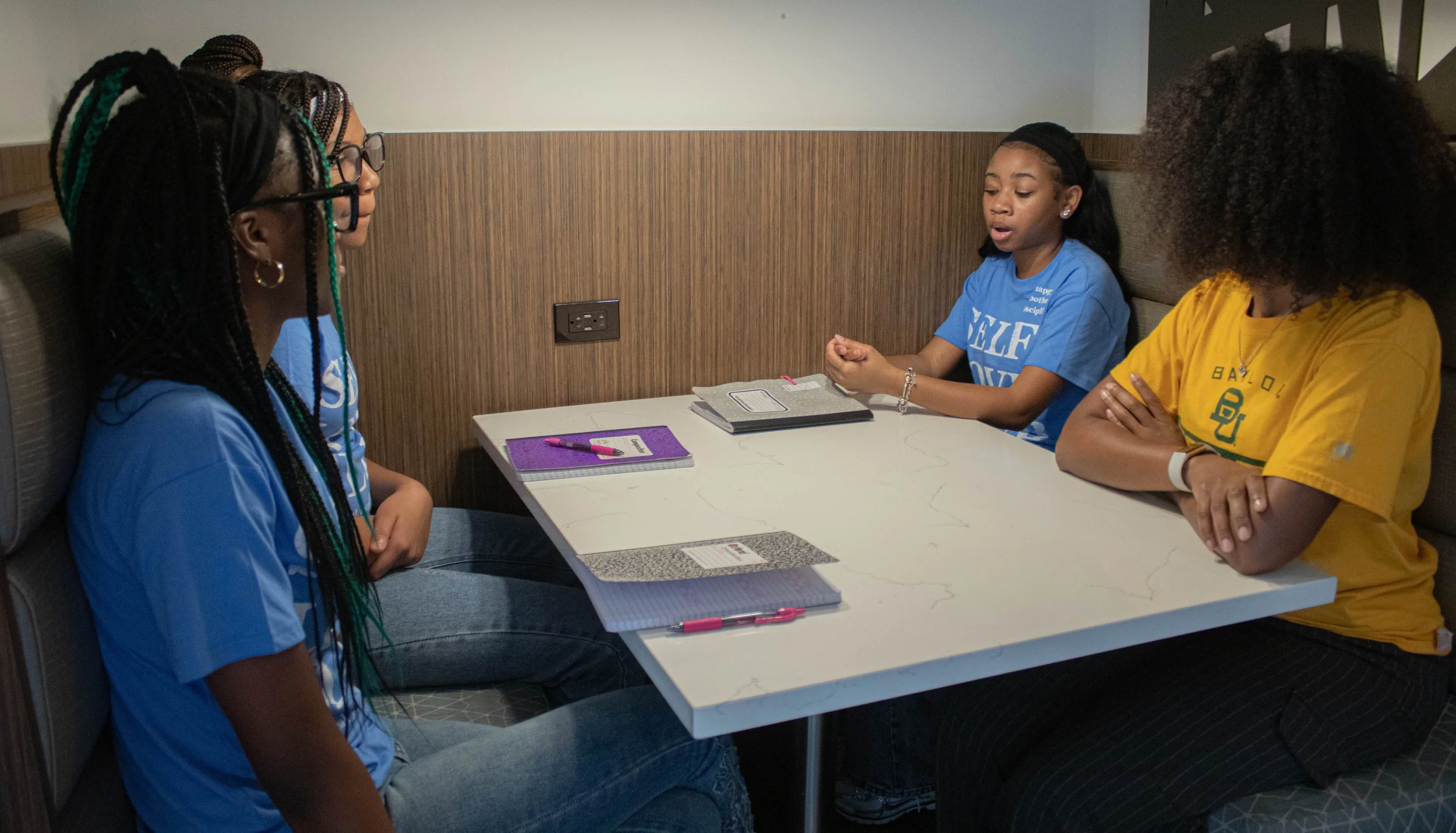 Four young women sitting around a table in a small booth, engaged in conversation, with notebooks and pens on the table.