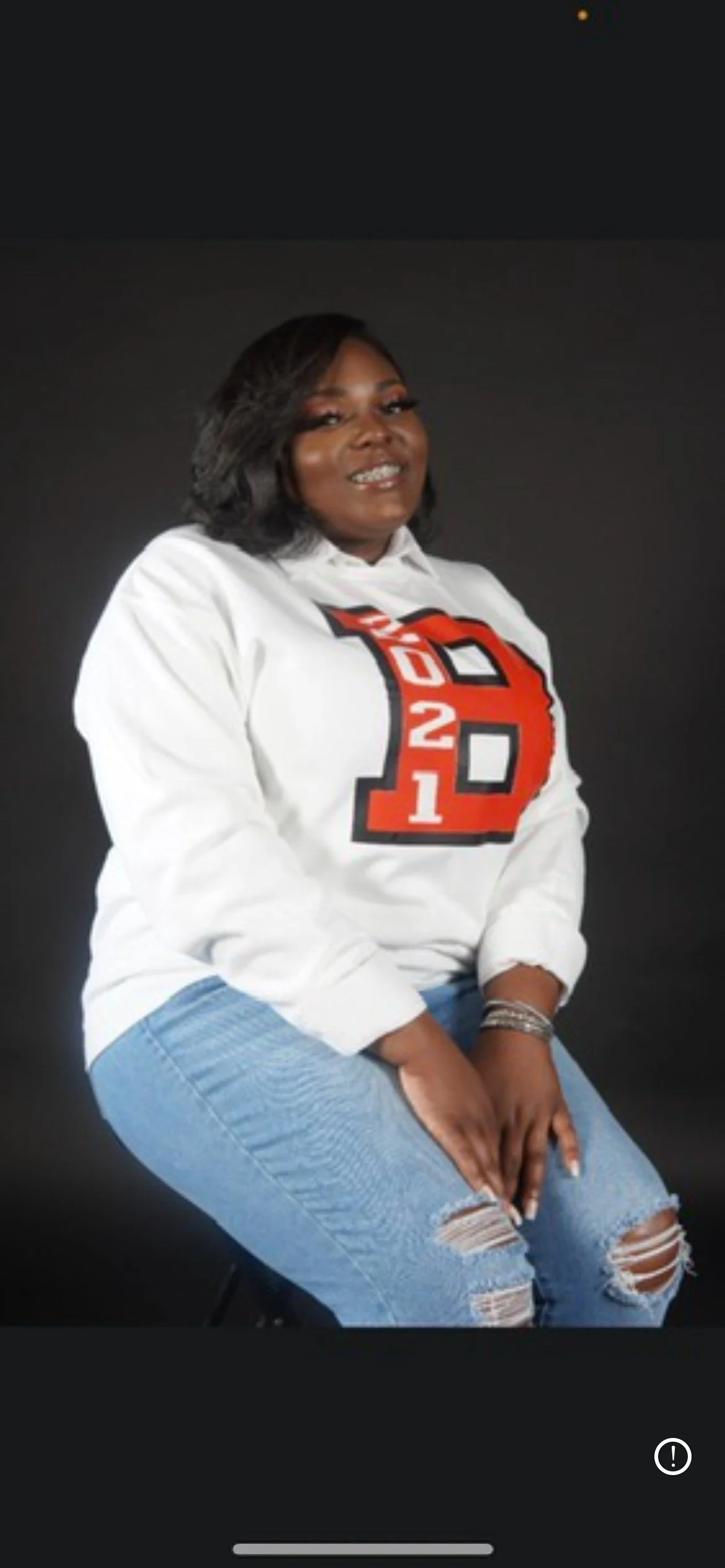 A woman with shoulder-length dark hair, wearing a white sweatshirt with red and black lettering, and ripped blue jeans, sitting against a black background.