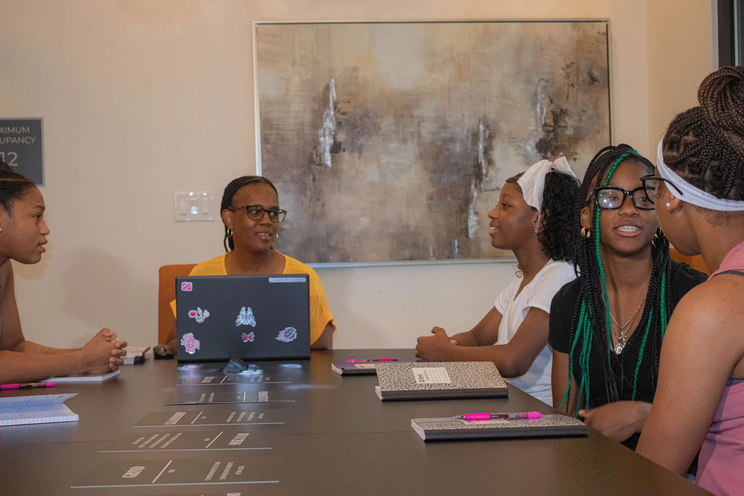 A diverse group of women sitting around a conference table, engaged in a discussion in a professional setting. There are notebooks, pens, and a laptop with stickers on it on the table. A large abstract painting is on the wall behind them.