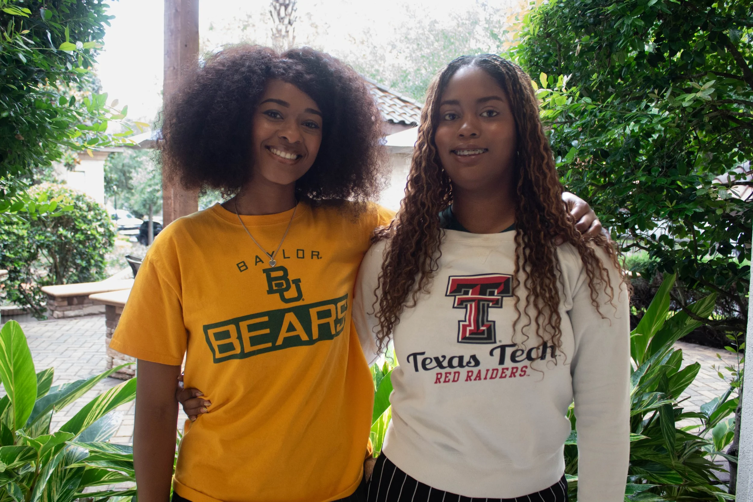 Two young women standing outdoors among greenery, smiling at the camera. One is wearing a yellow shirt with 'Baylor Bears' on it, and the other is wearing a white shirt with 'Texas Tech Red Raiders' on it.