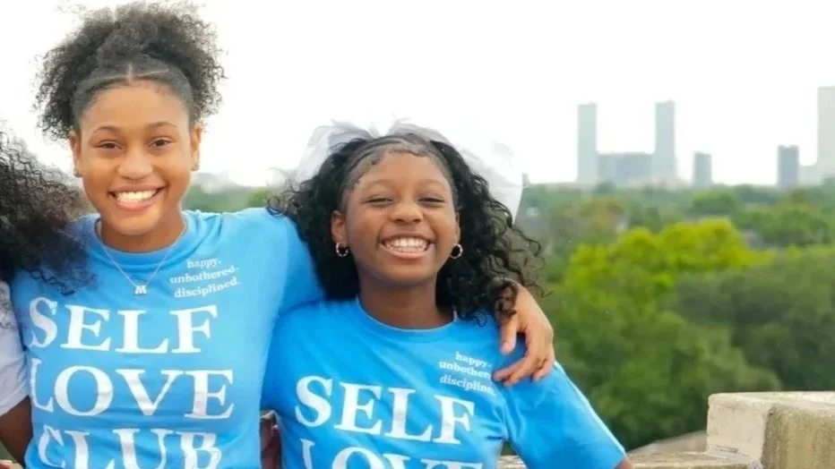 Two young girls smiling and wearing matching blue T-shirts with the words 'SELF LOVE CLUB' printed on them, standing outdoors with a cityscape and green trees in the background.