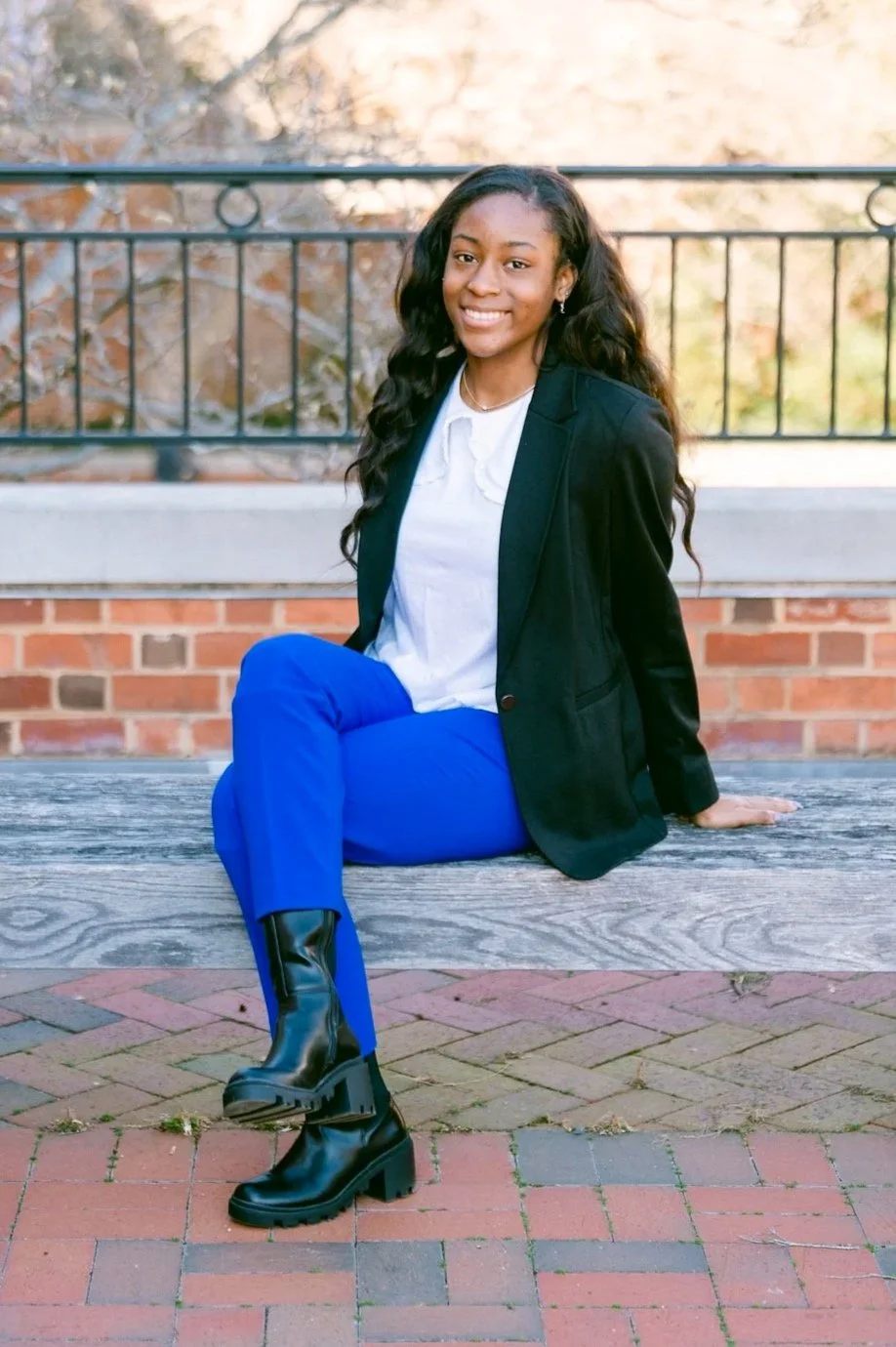 A young woman sitting on a wooden bench outdoors with a brick wall and metal railing behind her. She has long, wavy black hair and is smiling at the camera. She is dressed in a black blazer, white blouse, bright blue pants, and black chunky-heeled boots.