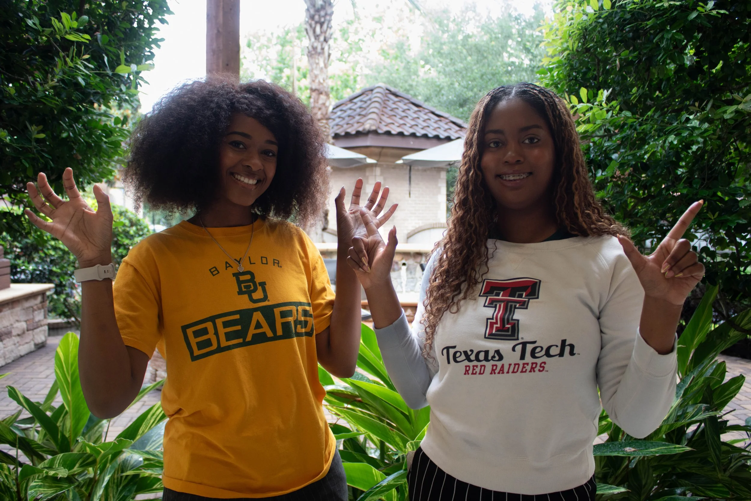 Two smiling women with curly hair standing outdoors among green plants, waving at the camera. One is wearing a yellow Baylor Bears t-shirt and the other a white Texas Tech Red Raiders sweatshirt.