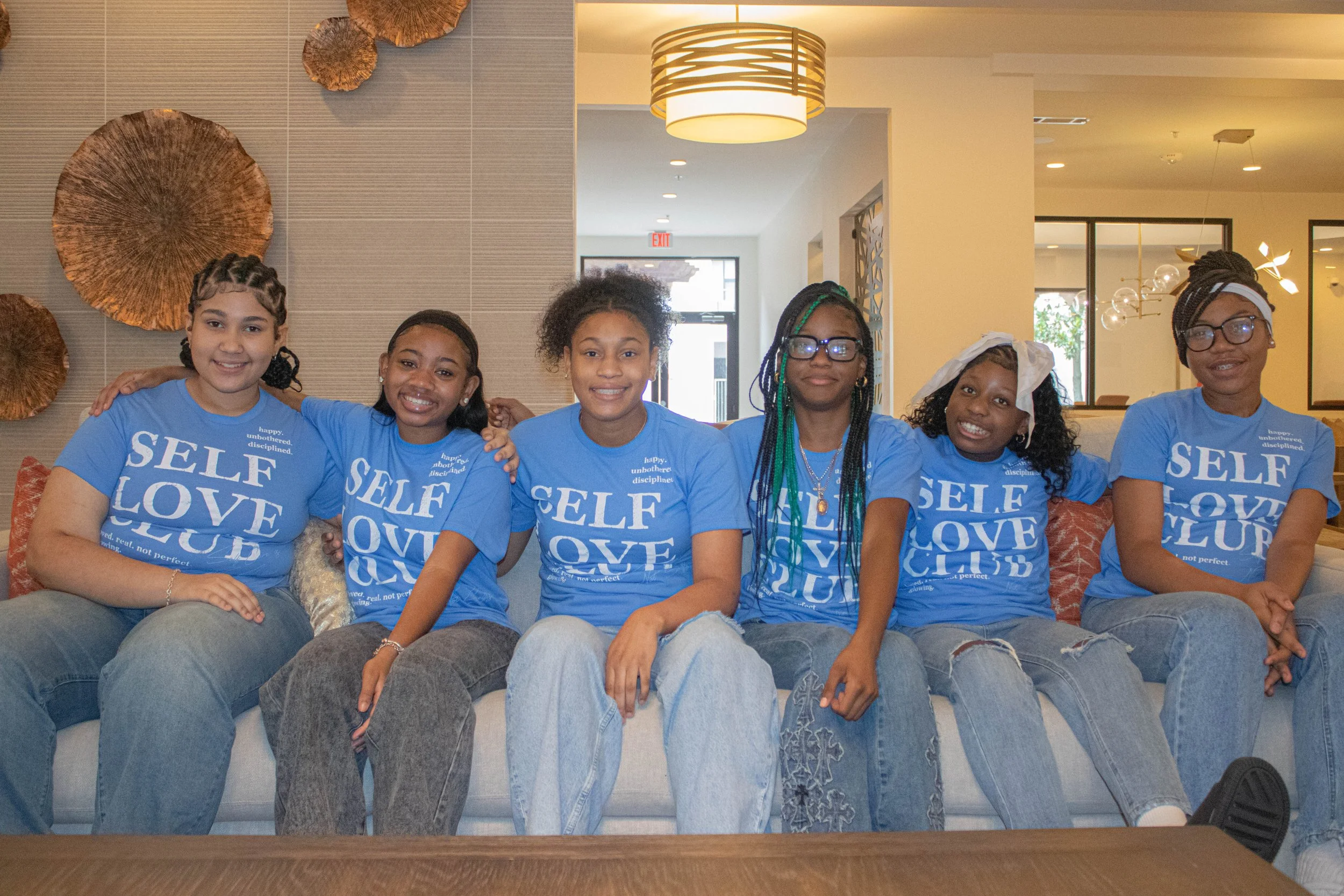 Six young women sitting on a couch in a modern, well-lit room, wearing matching blue T-shirts with the words 'SELF LOVE CLUB' printed on them, smiling and posing for the photo.