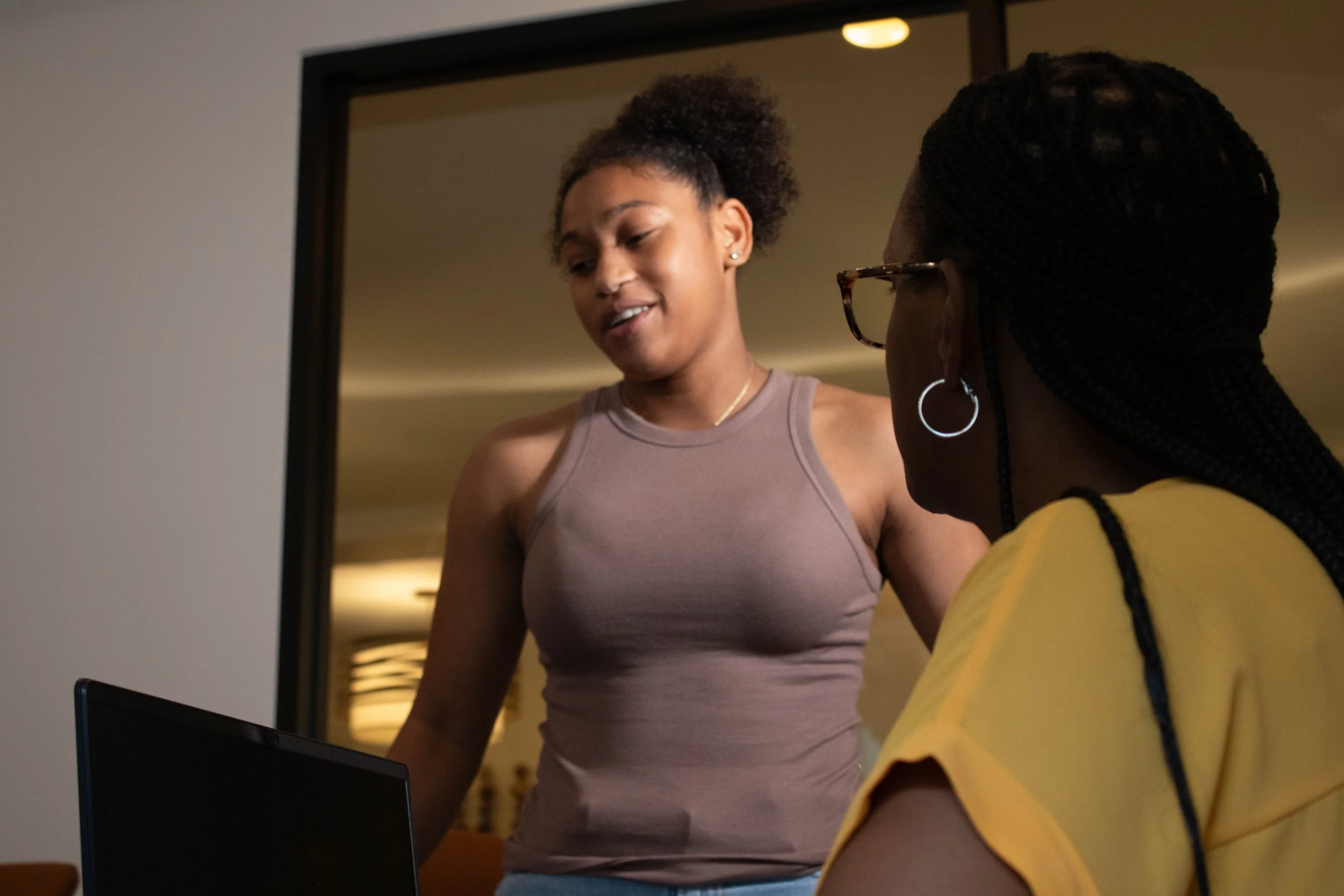 Two women looking at a computer screen, one wearing a lavender athletic top and the other wearing a yellow shirt with black straps, in an indoor setting with warm lighting.