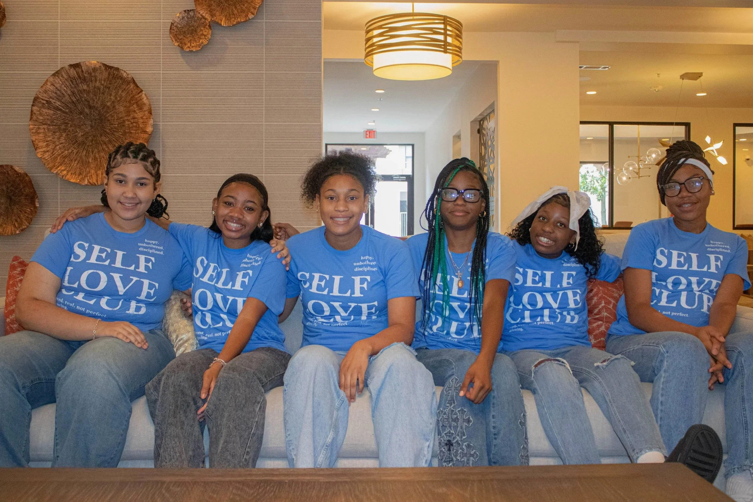 Six diverse women sitting closely together on a sofa, smiling, and wearing matching blue T-shirts with the words 'SELF LOVE CLUB' printed in white.