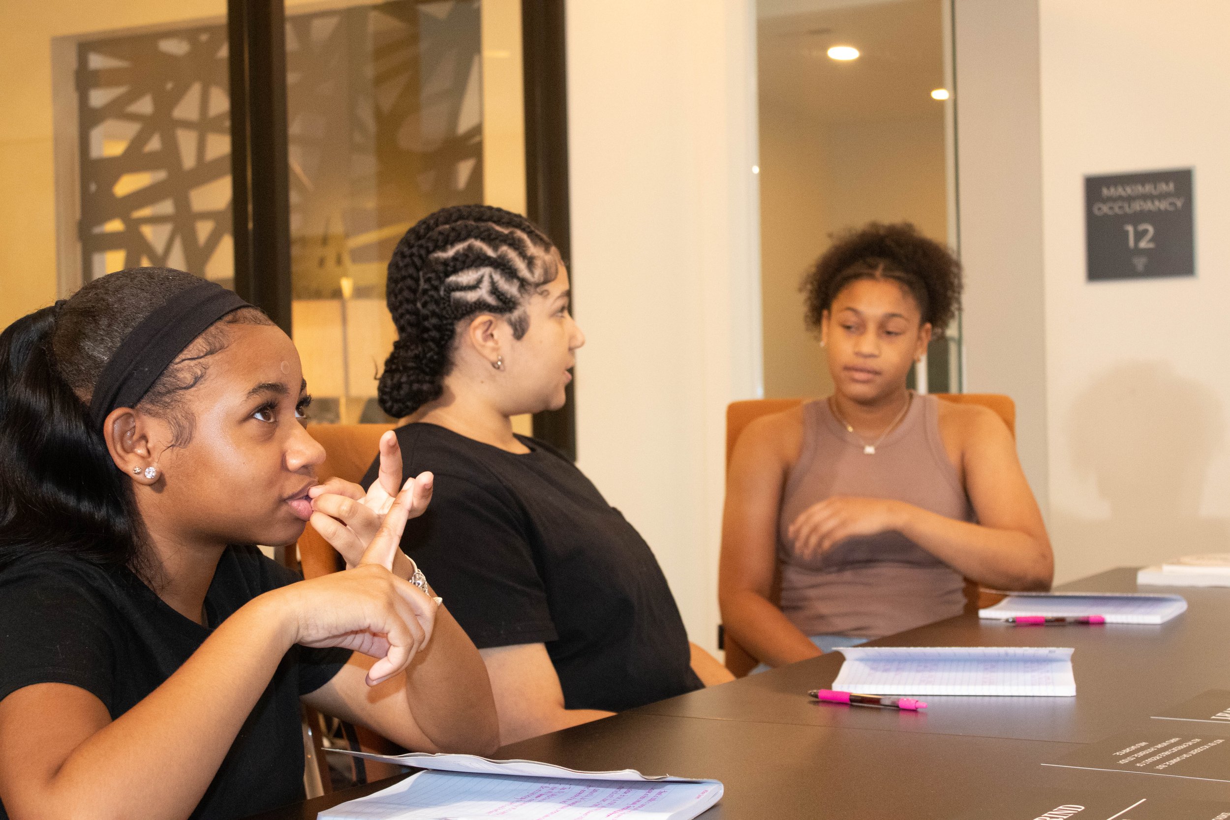 Three women are seated around a table, engaged in a discussion or meeting. They have notebooks and pens in front of them in a well-lit room with glass walls and a sign that reads 'Maximum Occupancy 12'.