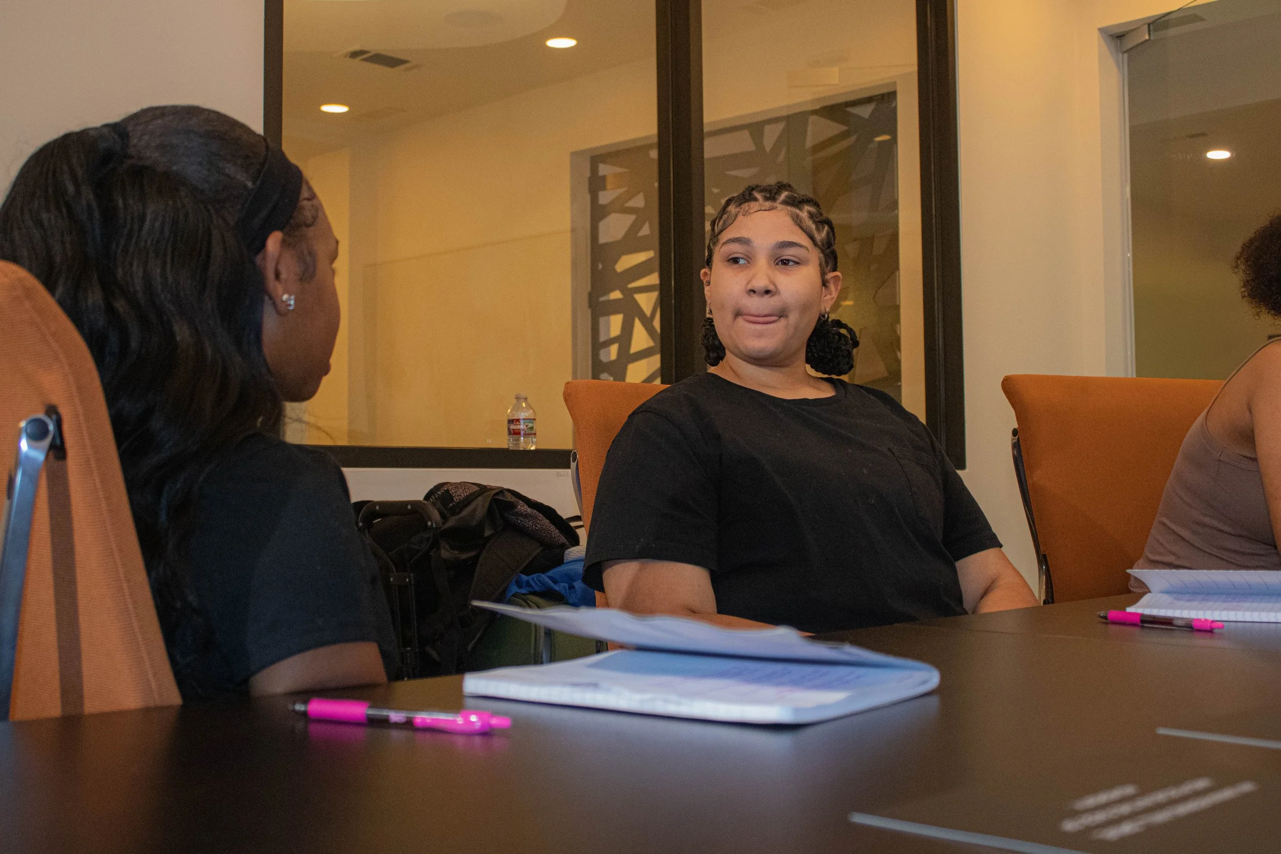 A young girl with braided hair and earrings sitting at a table during a discussion, with a woman with dark hair and earrings facing her. The table has notebooks, pens, and a water bottle, in a modern room with glass partitions and warm lighting.