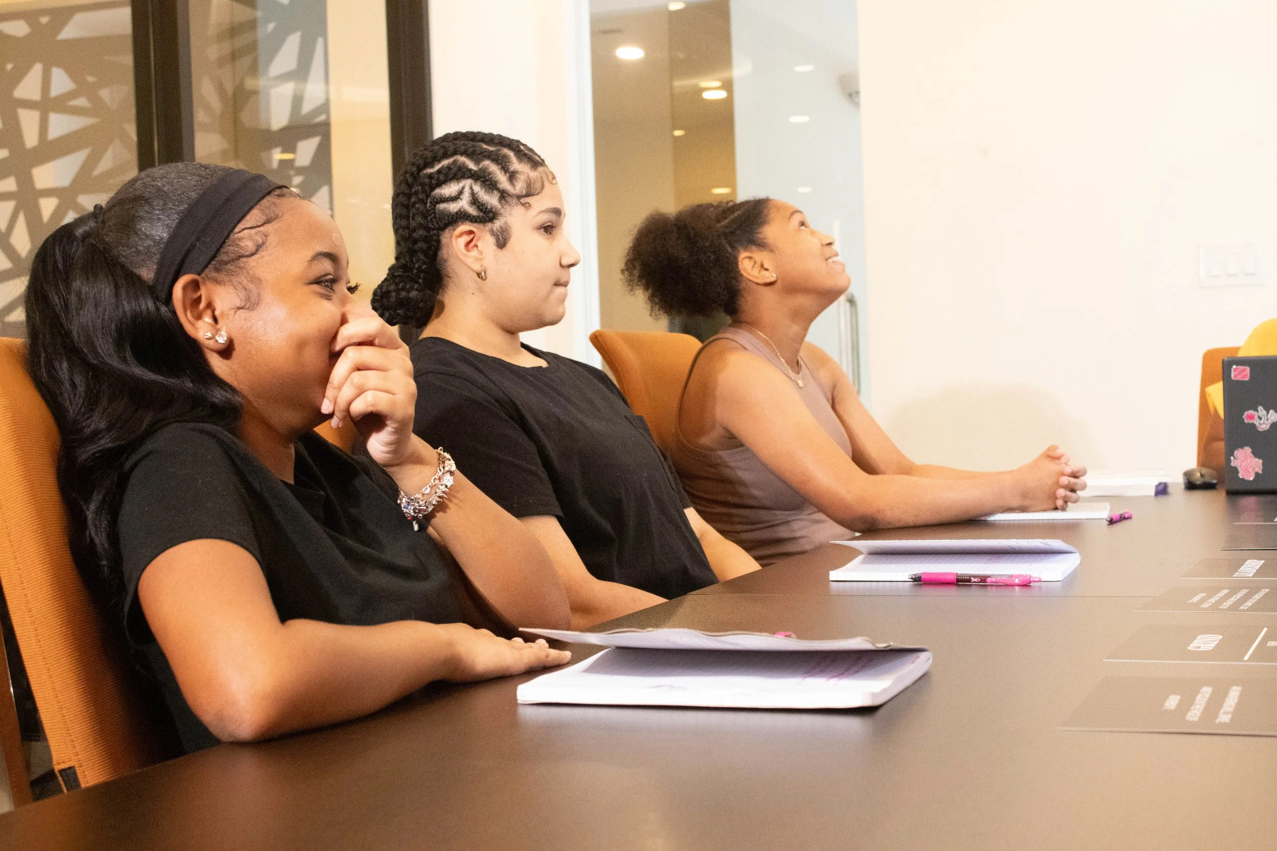 Three women sitting at a conference table, attentively listening. They have notebooks, pens, and papers in front of them. The woman on the right is smiling and looking upwards, the woman in the middle has tatoos and her hair in braids, and the woman 