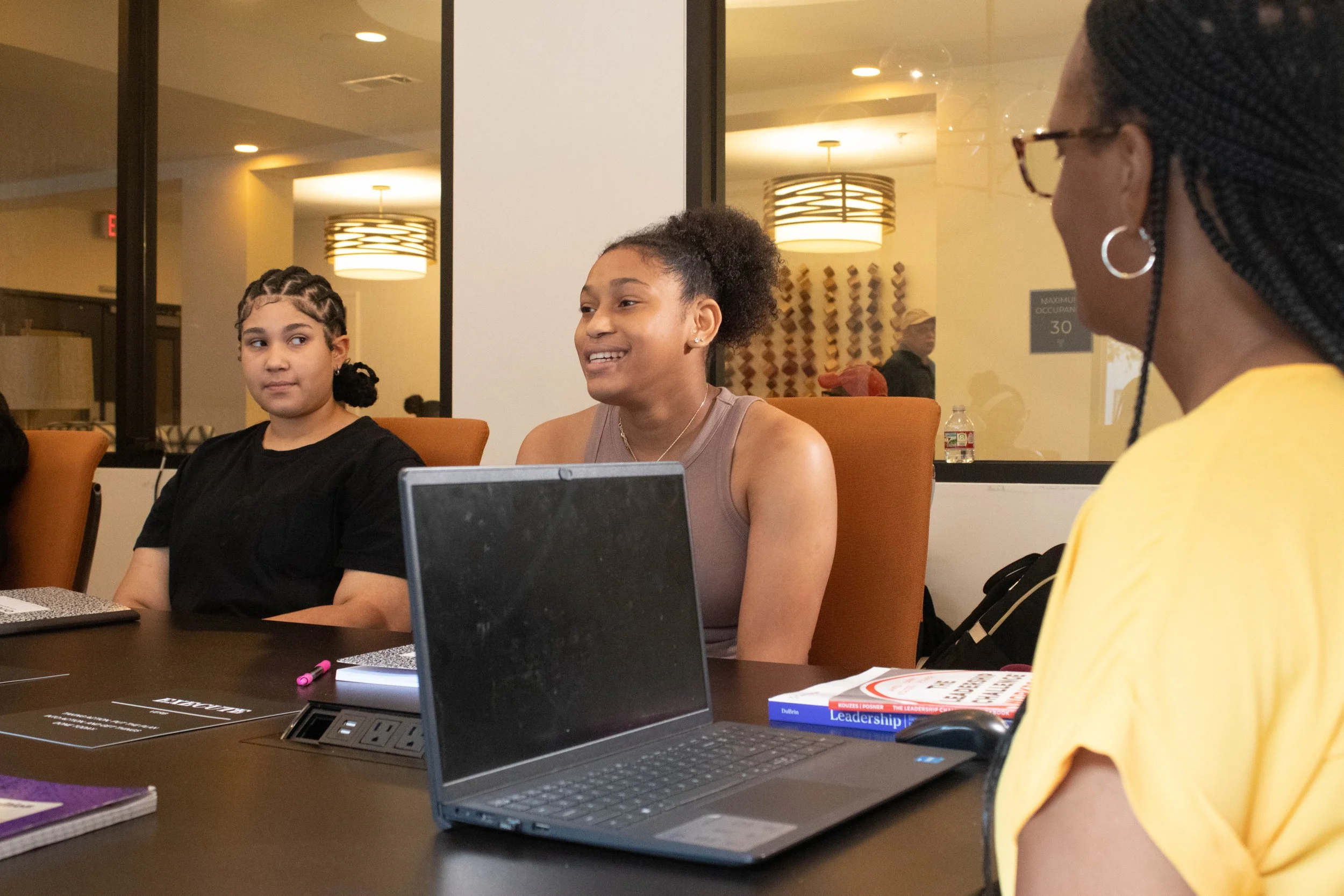 Three women seated at a conference table in a meeting room, engaging in conversation. One woman with dark braids and earrings in a yellow shirt is turned to the side, the woman with curly hair and a brown top is smiling and speaking, and the third wo