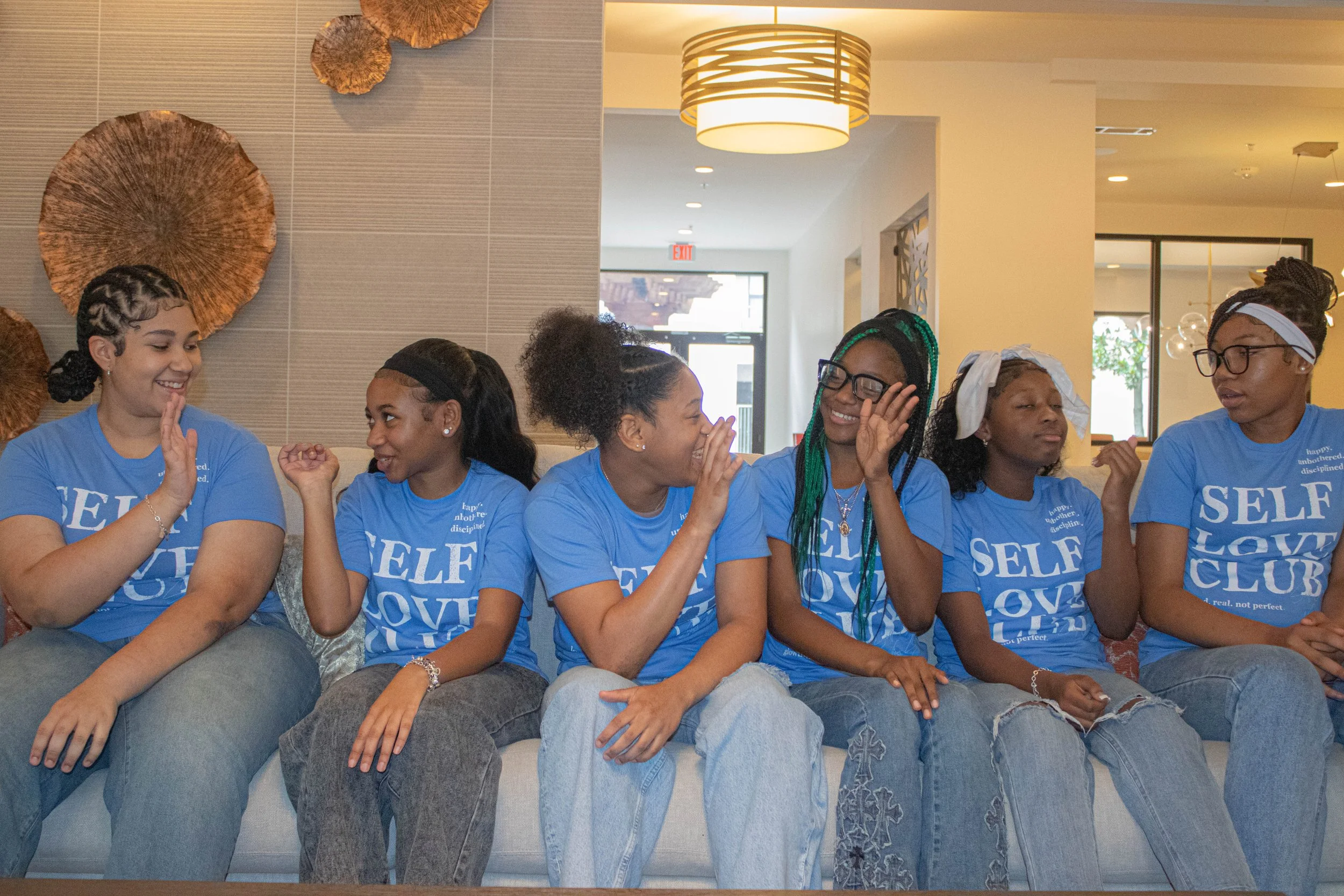 A group of seven women sitting together on a couch, wearing matching blue t-shirts that say 'SELF LOVE CLUB,' smiling, laughing, and engaging with each other in a casual setting inside a well-lit room.