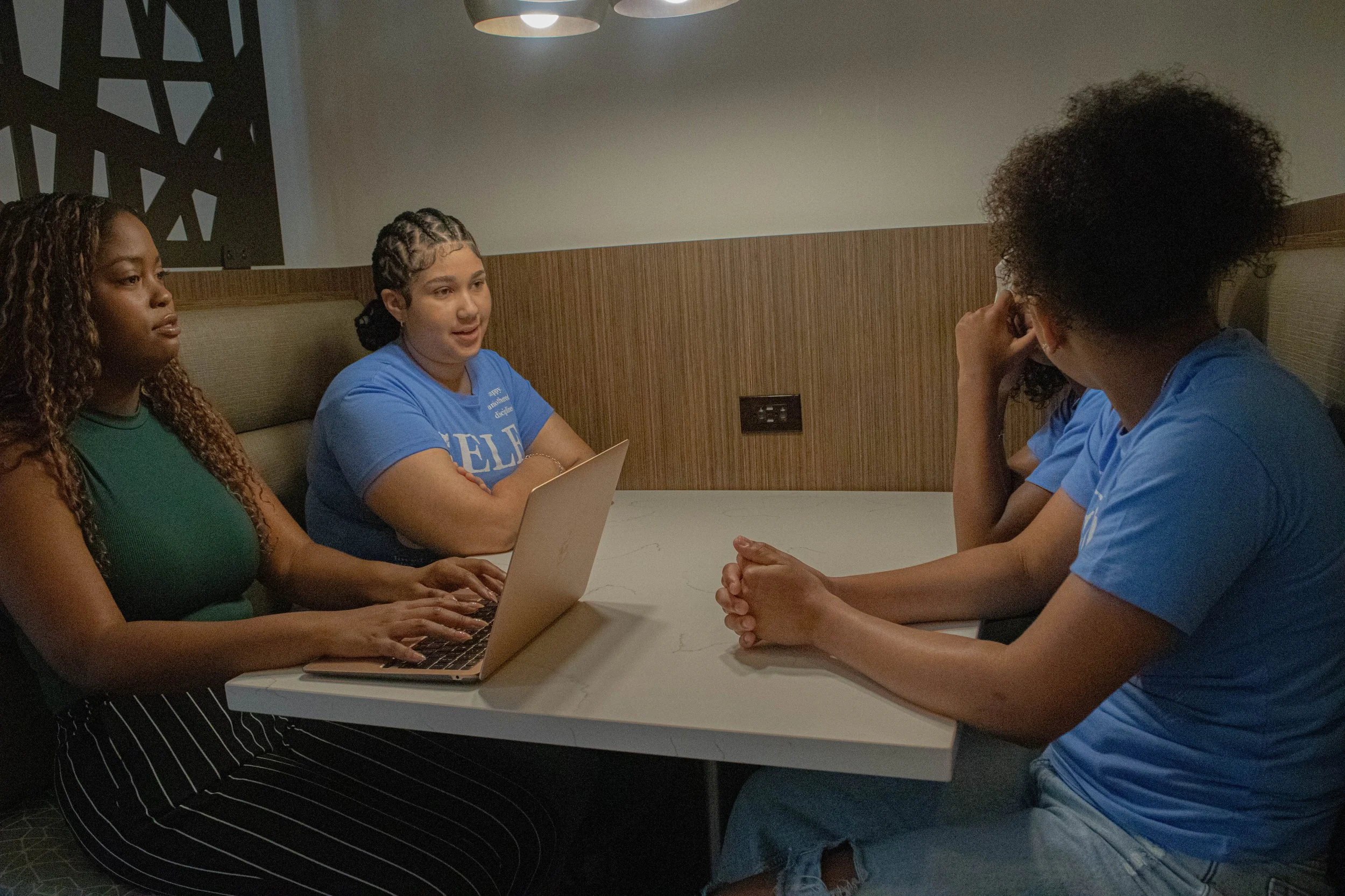 A group of five women sitting around a table having a discussion. One woman is typing on a laptop, while another is speaking, and the others are listening attentively, in a room with a wooden wall and hanging lights.