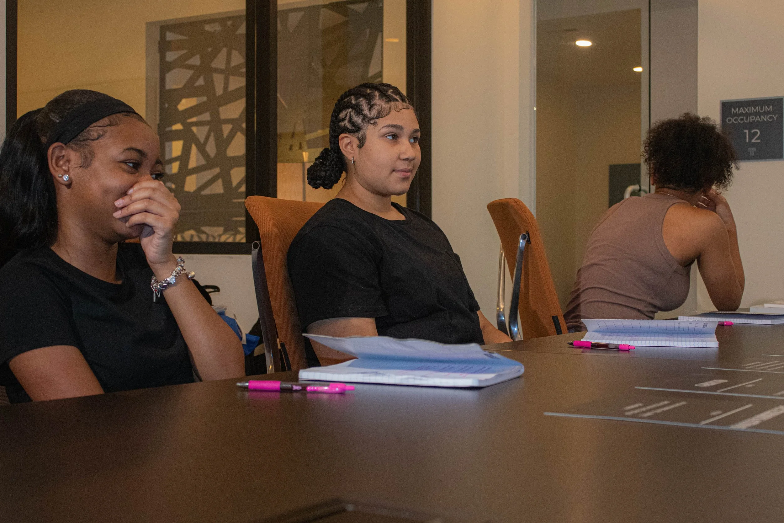 Three young women sitting at a table with notebooks and pens, engaged in a meeting or class inside a modern conference room.