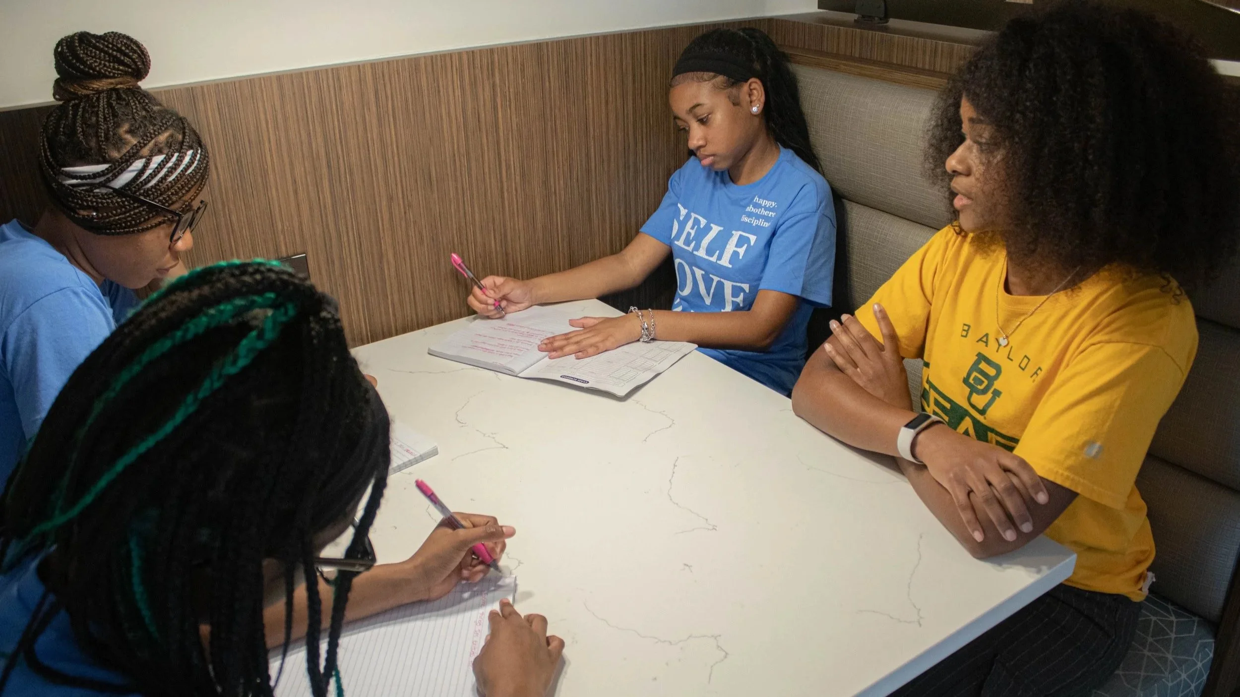 Group of young women sitting at a table, some writing in notebooks, in a casual indoor setting.