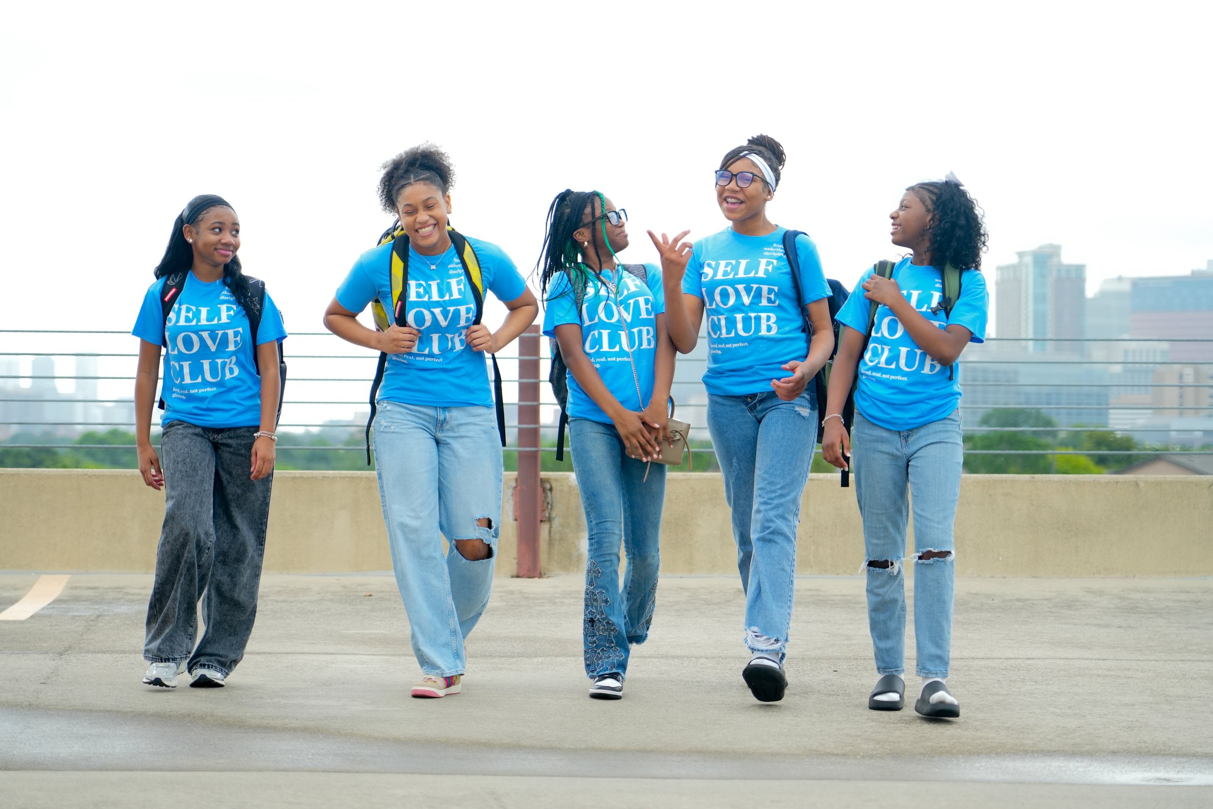 Five young African American girls walking and talking on a rooftop parking lot, wearing matching blue T-shirts that say 'Self Love Club,' with a city skyline in the background.