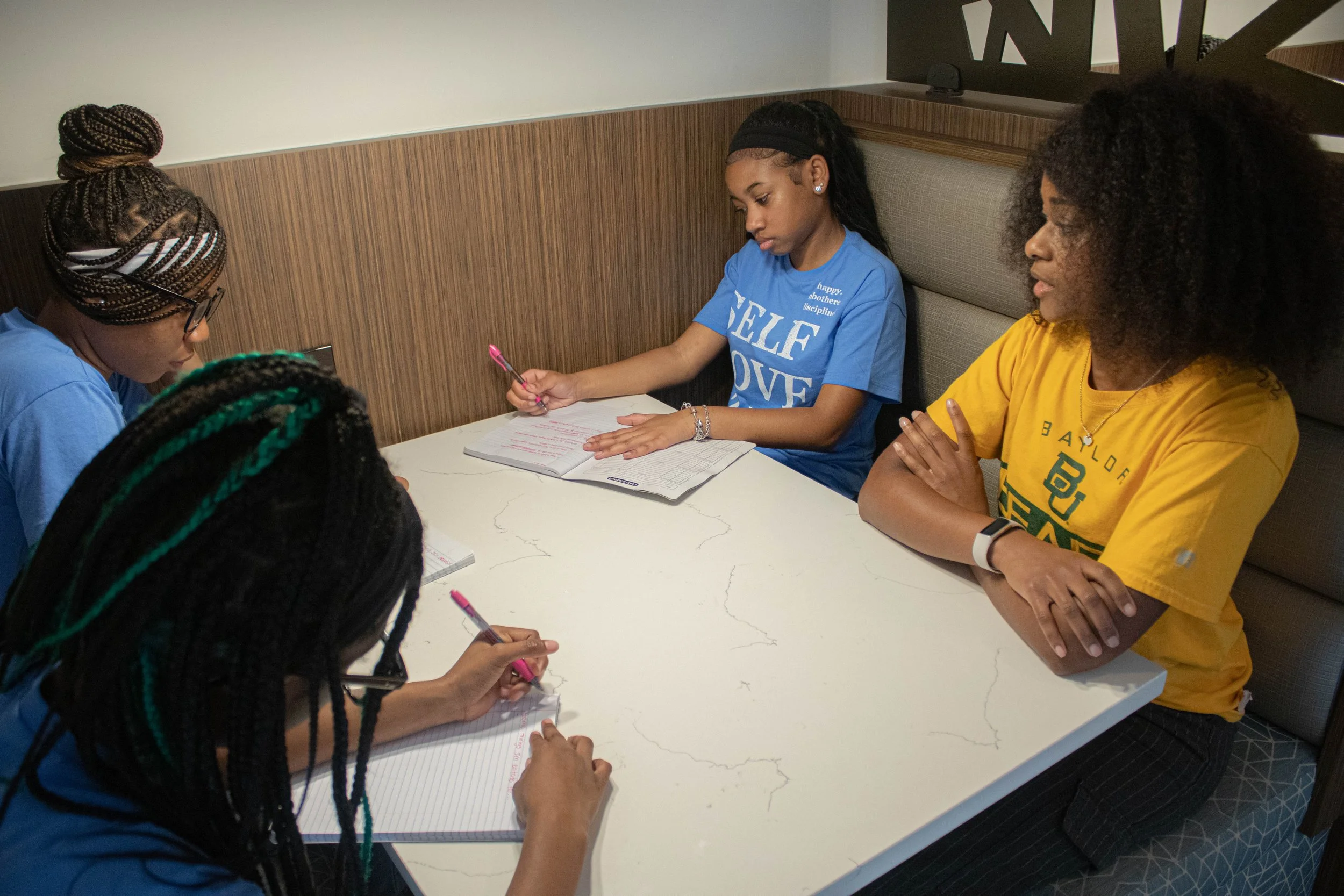 Four women sitting around a white table in a casual indoor setting. Two women are taking notes, while the other two observe. The woman in the blue shirt has long black hair and is writing, the woman in the yellow shirt has curly hair and is watching,