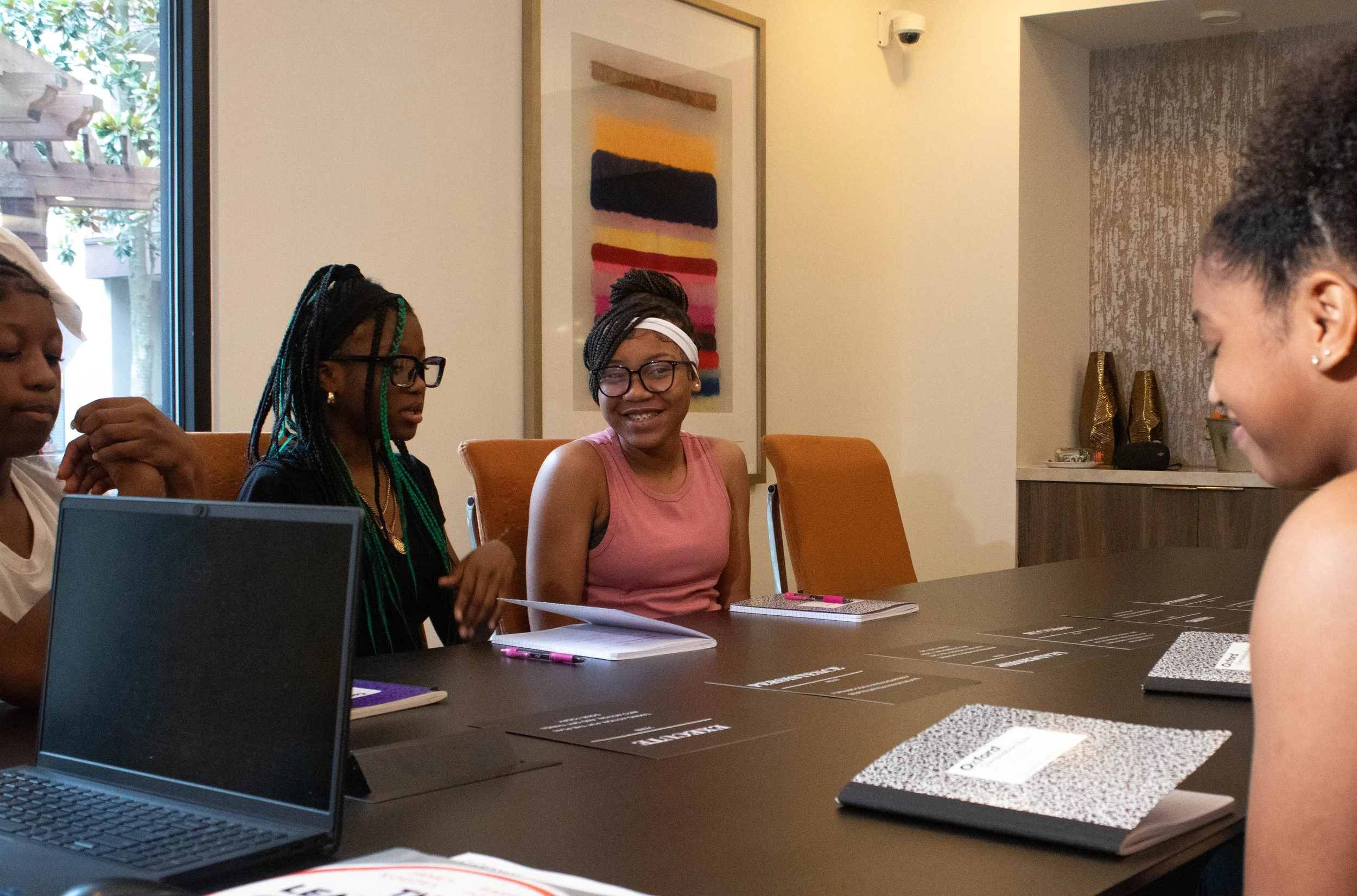 Four women sitting around a table in a meeting room, with notebooks, pens, and laptops in front of them. One woman is smiling, the others are listening or speaking.
