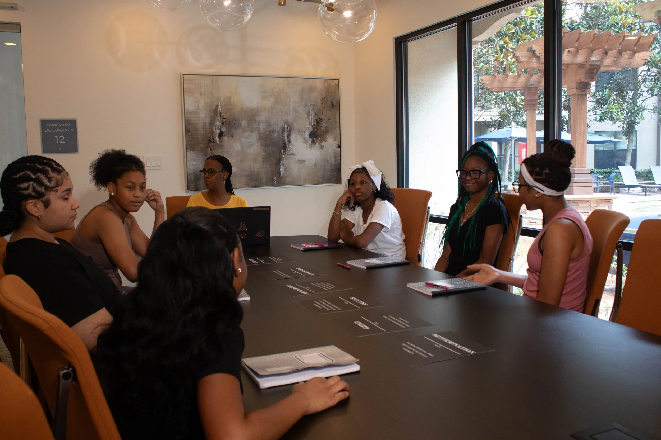 A diverse group of young girls sitting around a conference table in a well-lit room with large windows, engaged in a meeting or discussion.
