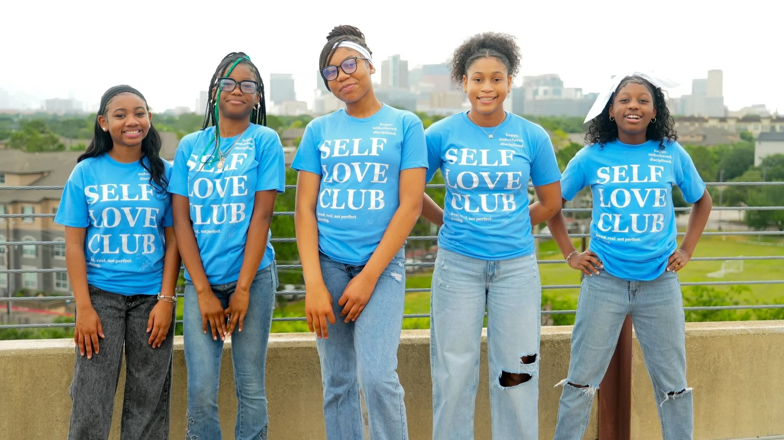 Five young girls standing outdoors, wearing matching blue T-shirts that read 'Self Love Club,' with a city skyline in the background.