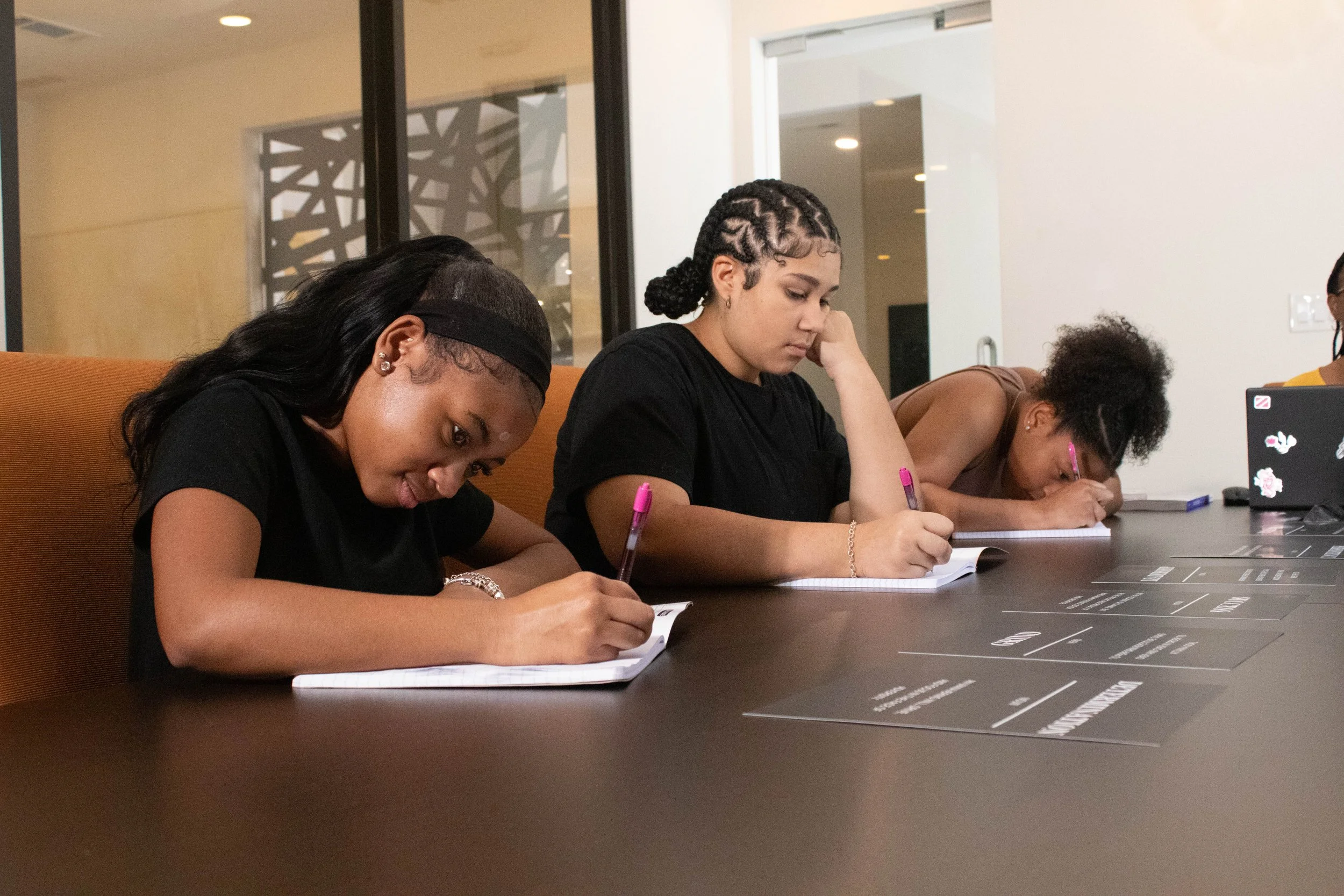 Four young women sitting at a table, writing in notebooks during a meeting or study session in a modern office or classroom.