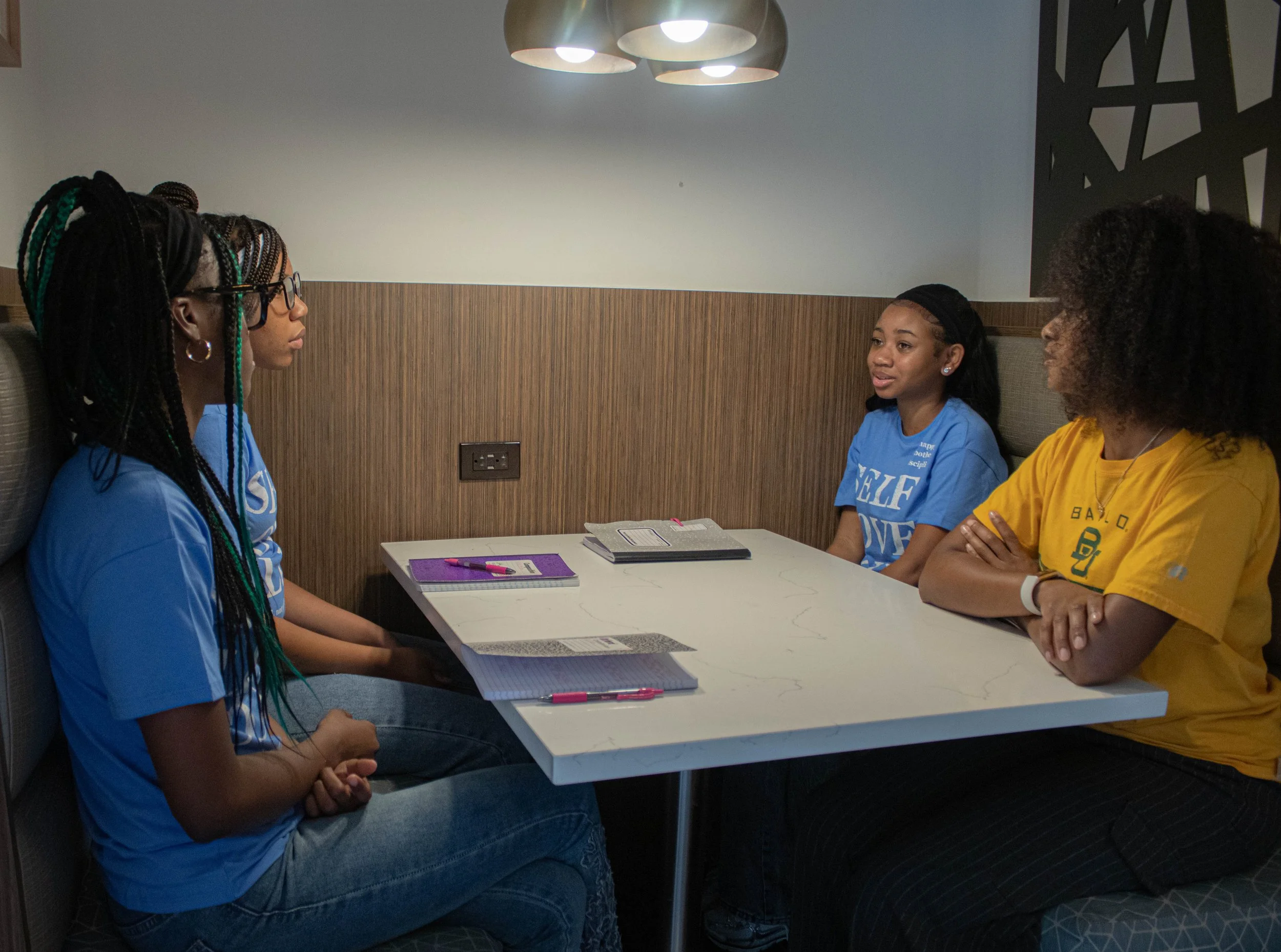 Four young women sitting at a table in a restaurant or cafe, having a conversation. They are dressed casually, and two of them are wearing blue shirts while one is in a yellow shirt. There are notebooks, pens, and a smartphone on the table.