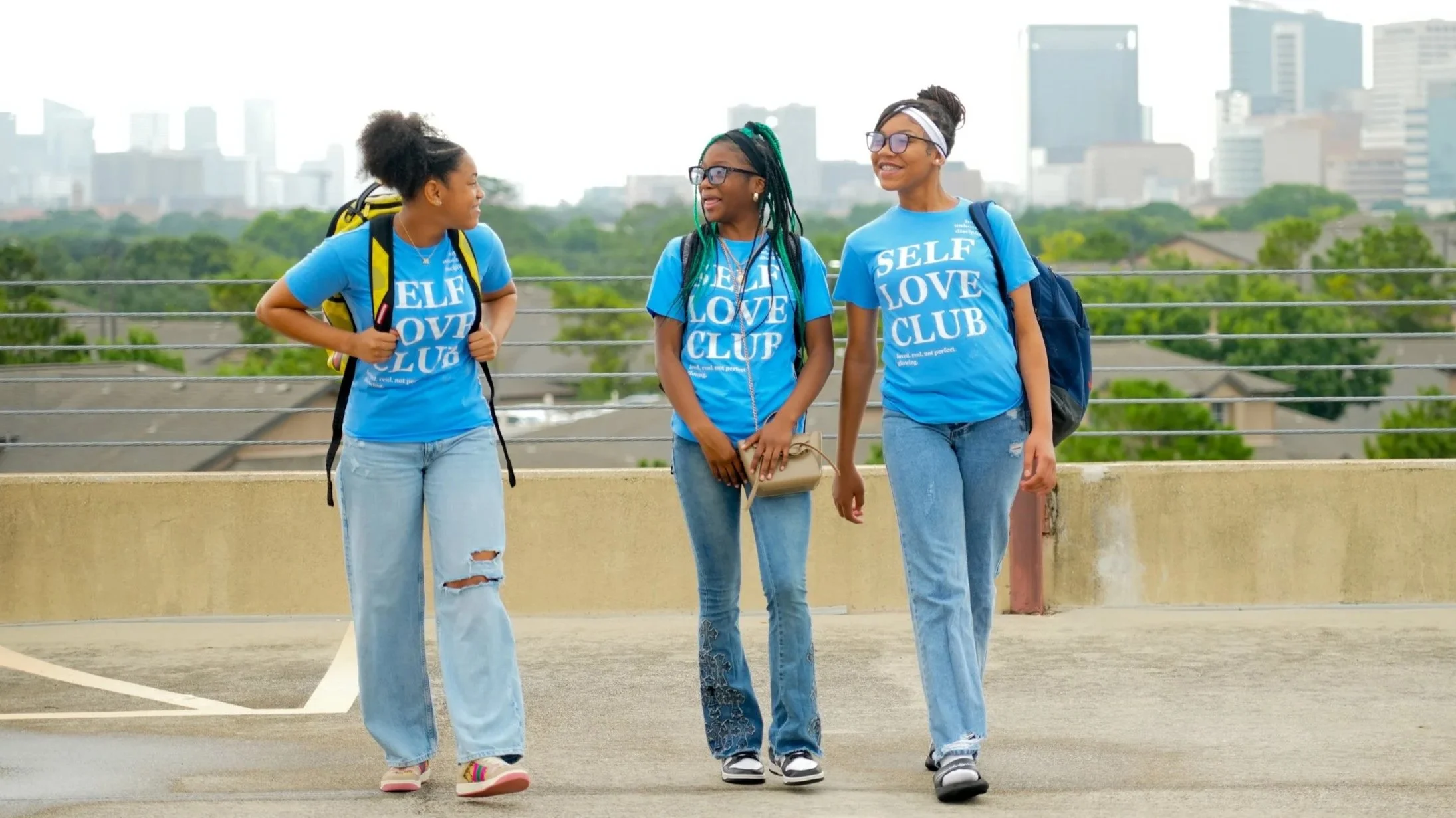 Three young women walking and talking on a rooftop, wearing matching blue t-shirts with the words 'SELF LOVE CLUB', with city buildings in the background.