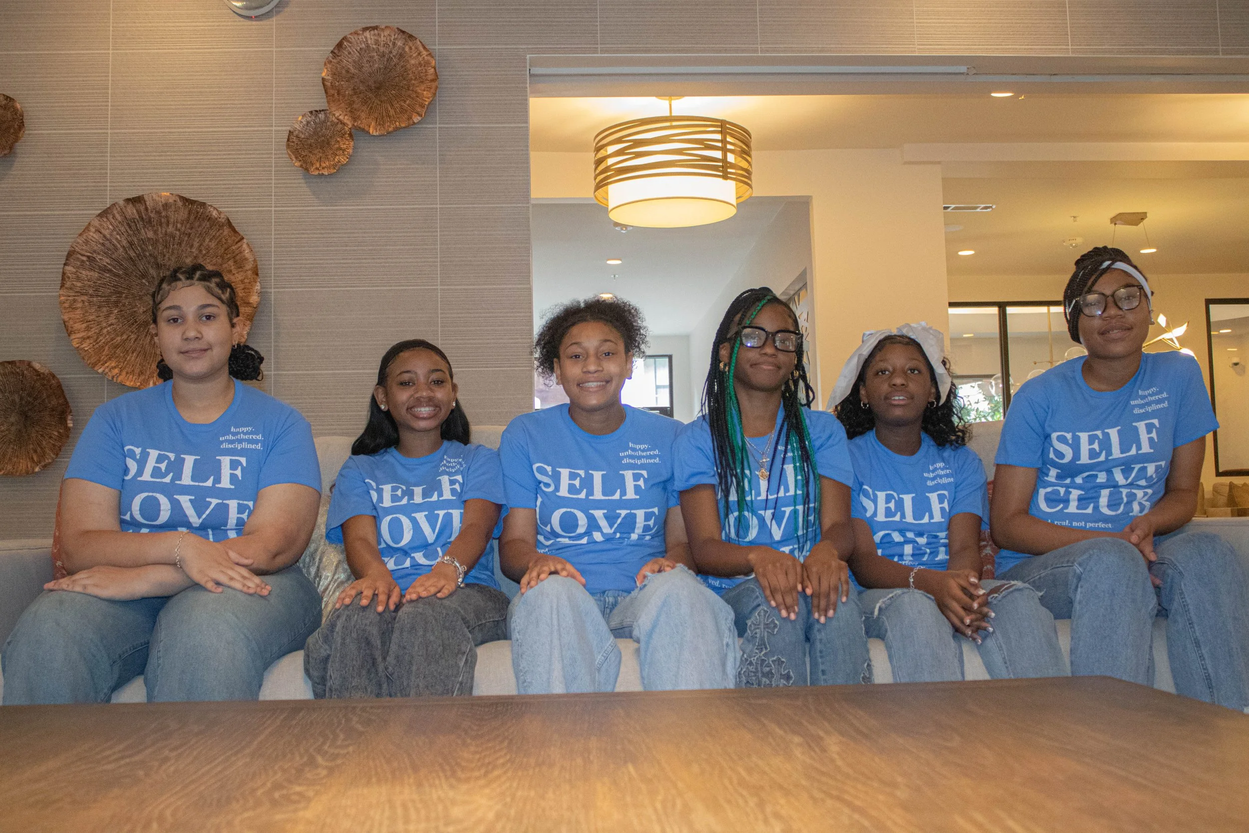 Six diverse young girls sitting side by side on a couch, wearing blue T-shirts with words about self-love, in a modern indoor setting with a decorative wall and ceiling lights.