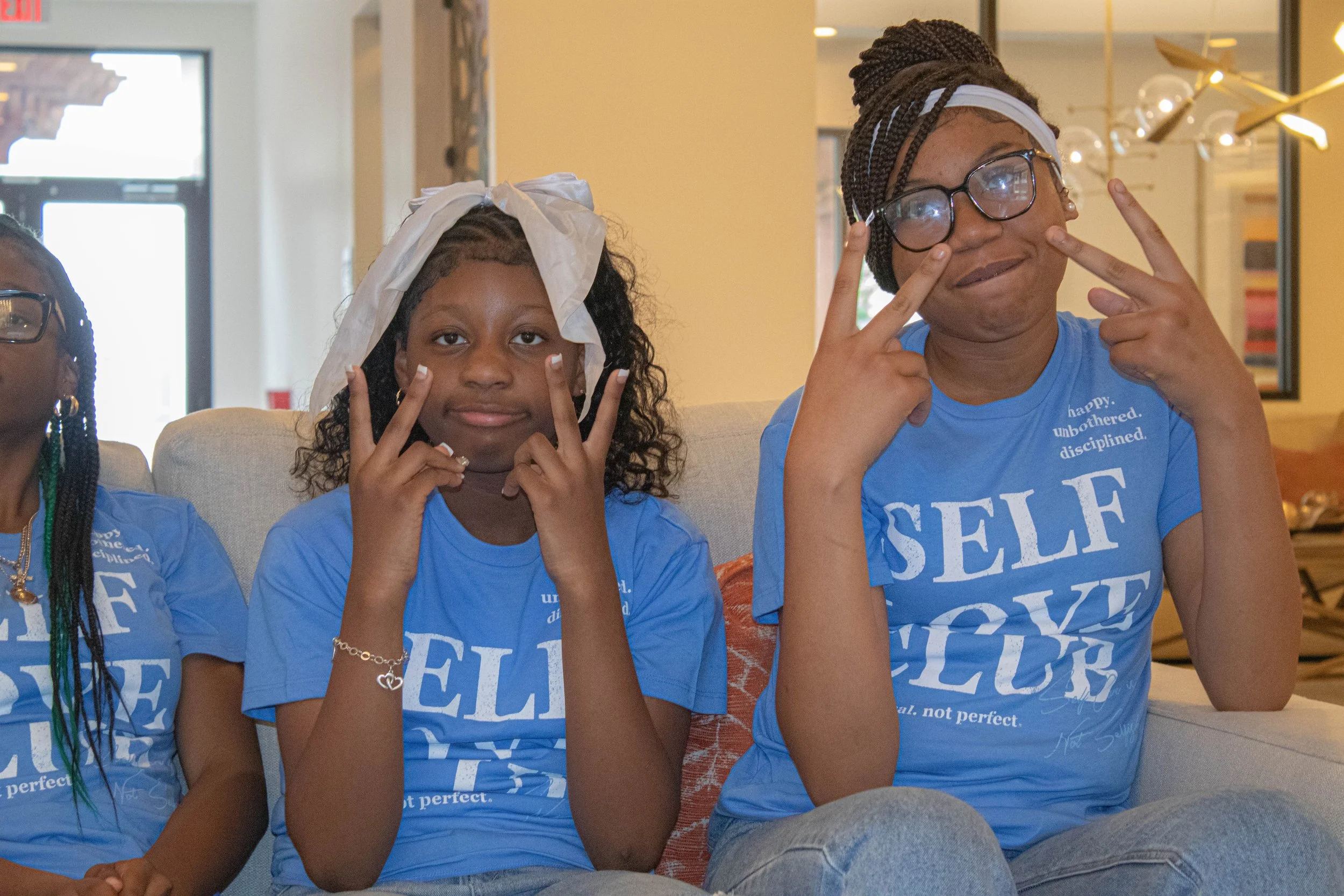 Two young women sitting indoors, both wearing blue t-shirts with white text. The woman on the left is making a peace sign with her fingers near her face, has curly hair covered with a white head wrap, and is accessorized with jewelry. The woman on th