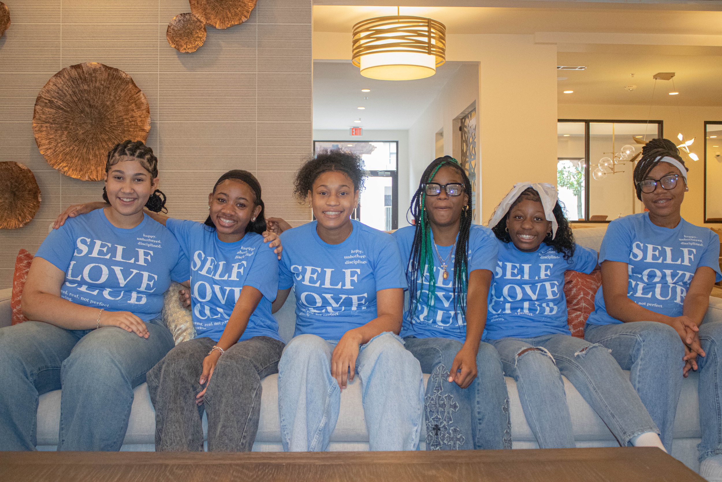 A group of seven smiling young girls with different hairstyles and glasses, sitting on a gray couch in a modern, well-lit indoor space, all wearing blue t-shirts with the words 'SELF LOVE' printed on them.