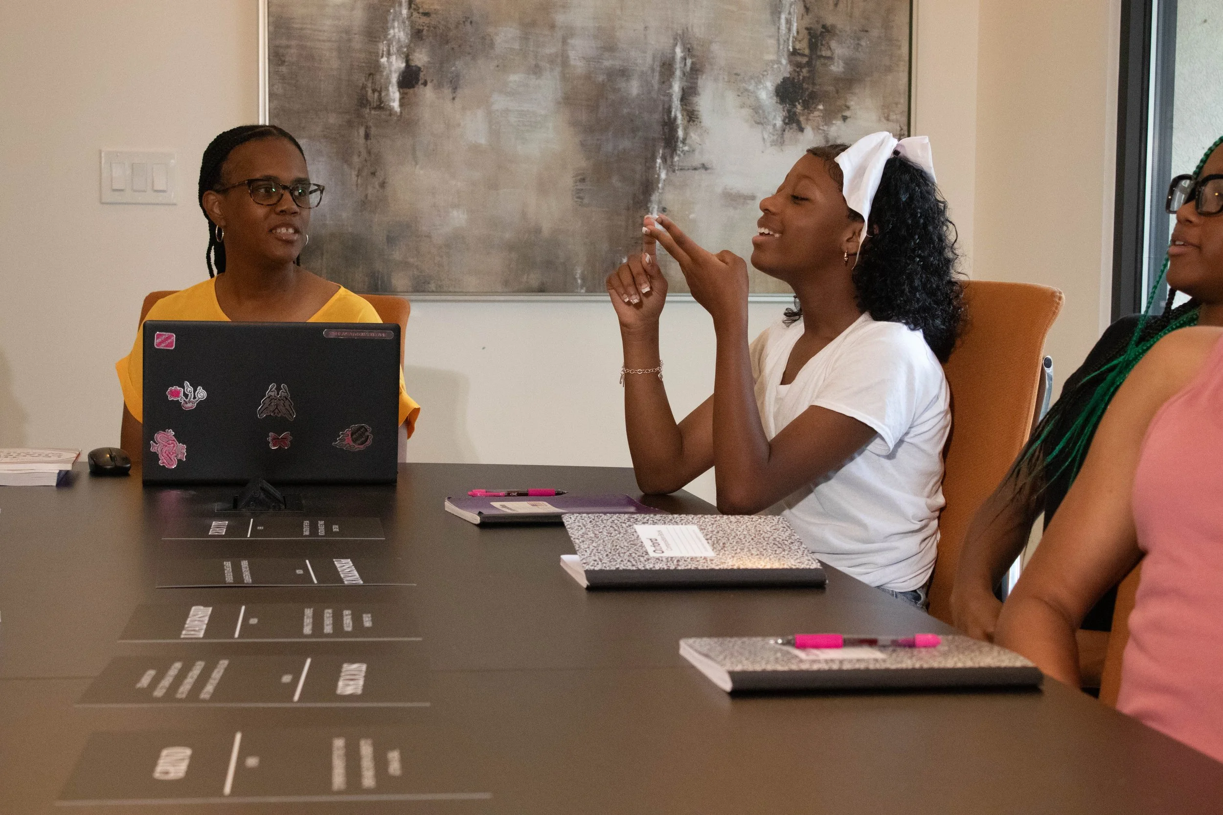 Three women sitting at a table in a meeting or discussion. One woman on the left is looking at the woman in the middle who is holding a pen and smiling. The woman in the middle has curly hair and a white headband. The third woman, on the right, is pa