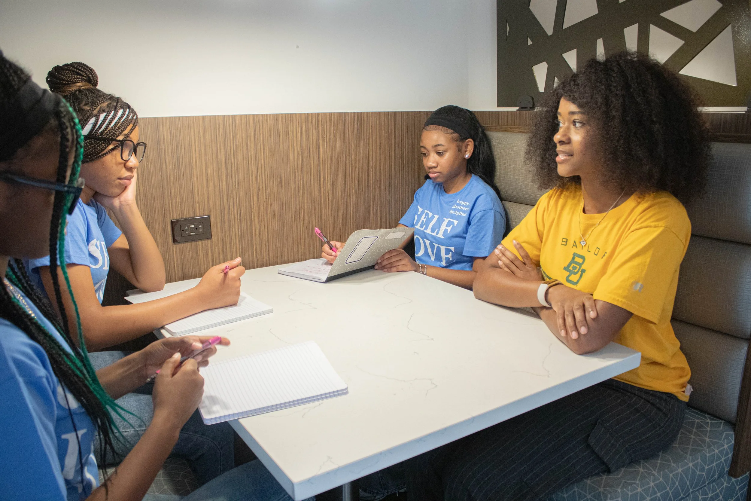 Group of four women sitting at a booth in a restaurant or cafe, engaged in a discussion or meeting, with notebooks and pens on the table.