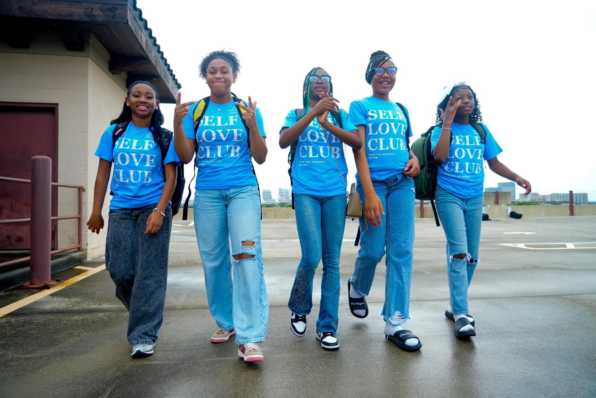 Five young girls standing together on a rooftop parking lot, wearing matching blue T-shirts that say 'Self Love Club,' with city buildings in the background.
