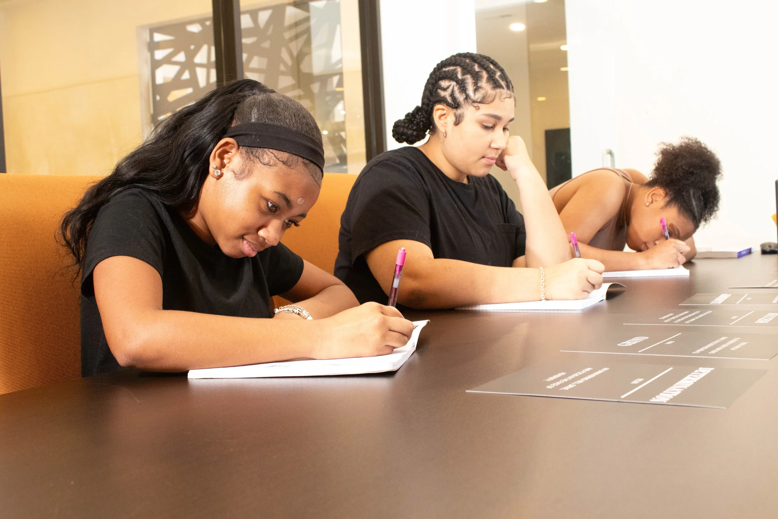 Three women sitting at a table, writing on notepads with pink pens, in an indoor setting with a window and decorative wall panel in the background.