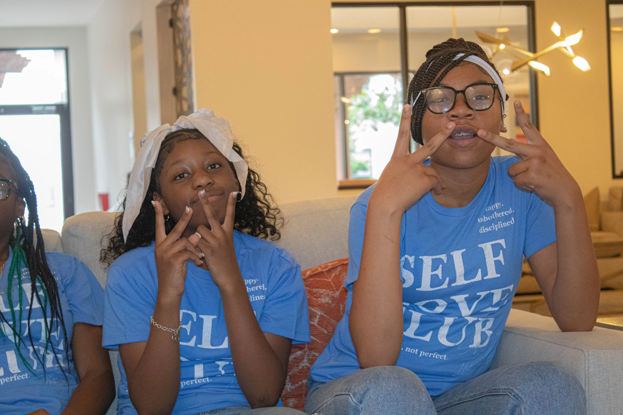 Two young women sitting on a couch making peace signs with their fingers and smiling at the camera in a bright indoor setting.