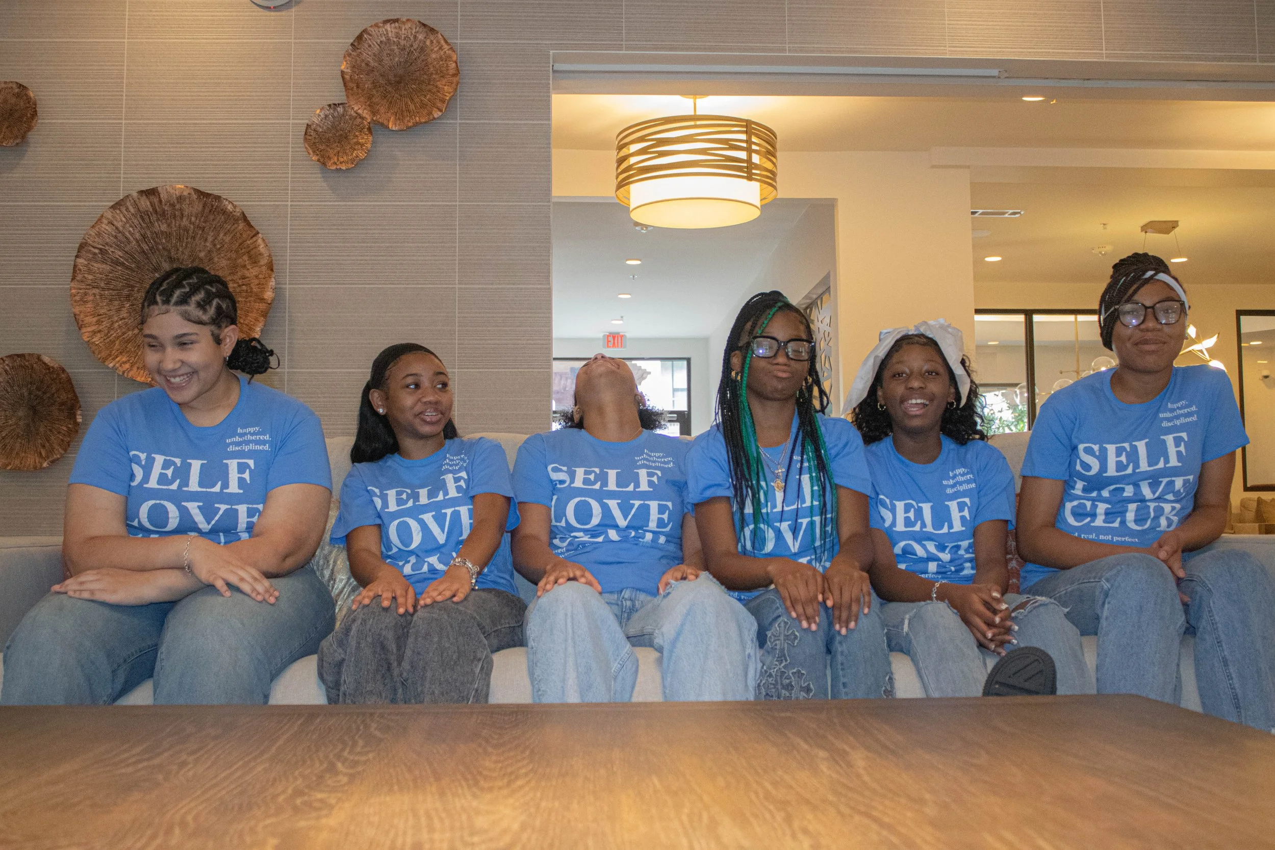 Six young women sitting side by side on a couch, all wearing matching blue t-shirts with the words 'Self Love Club' printed on them. They are smiling and appear to be at a social gathering or event in a well-lit, modern indoor space.