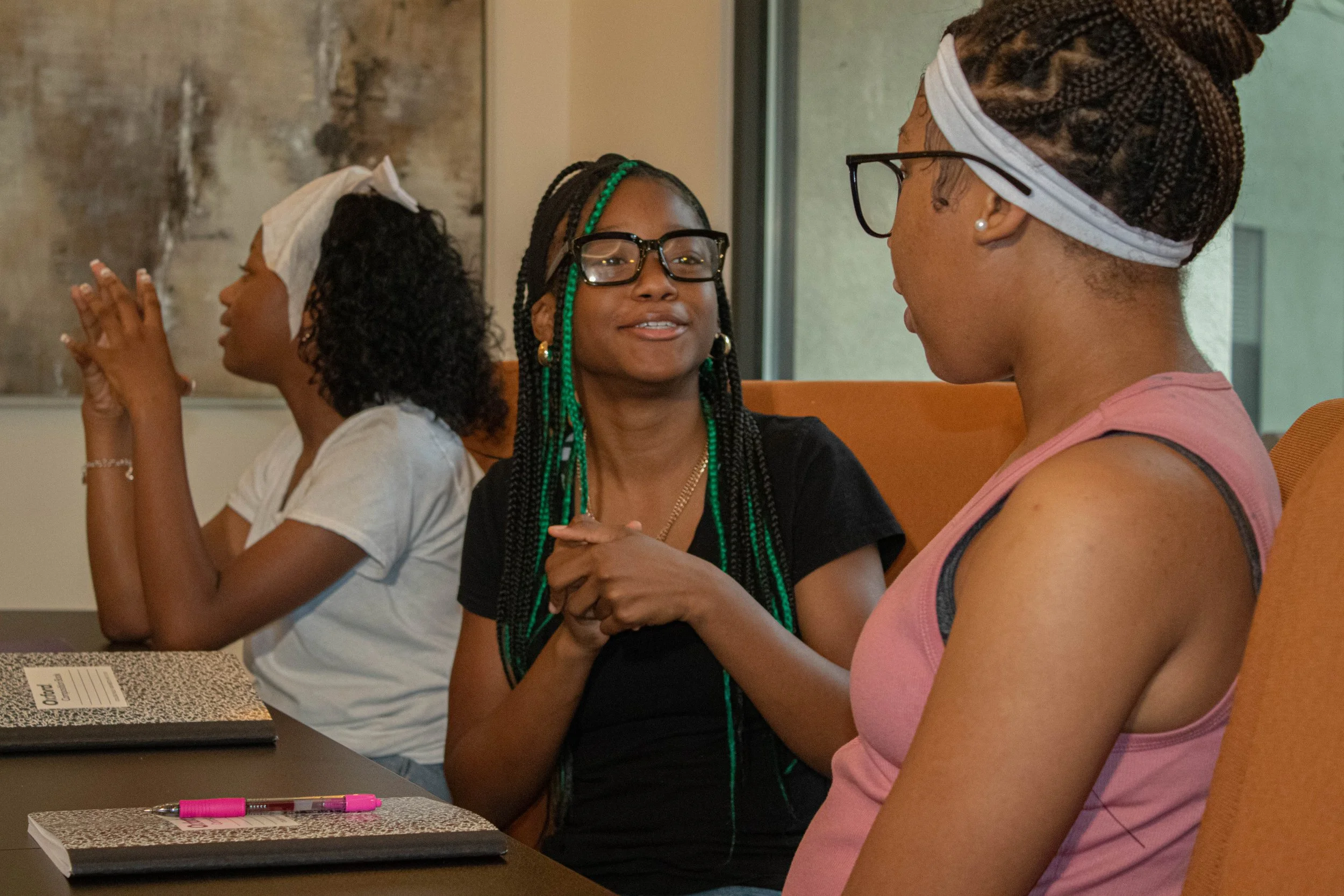 Three women sitting at a table talking, with two of them holding hands and one with hands clasped together in prayer. The woman on the right is wearing glasses, a pink tank top, and has her hair in braids covered with a scarf. The woman in the middle is wearing glasses and has long braids with green beads. The woman on the left has curly hair, a white headwrap, and is wearing a white shirt.