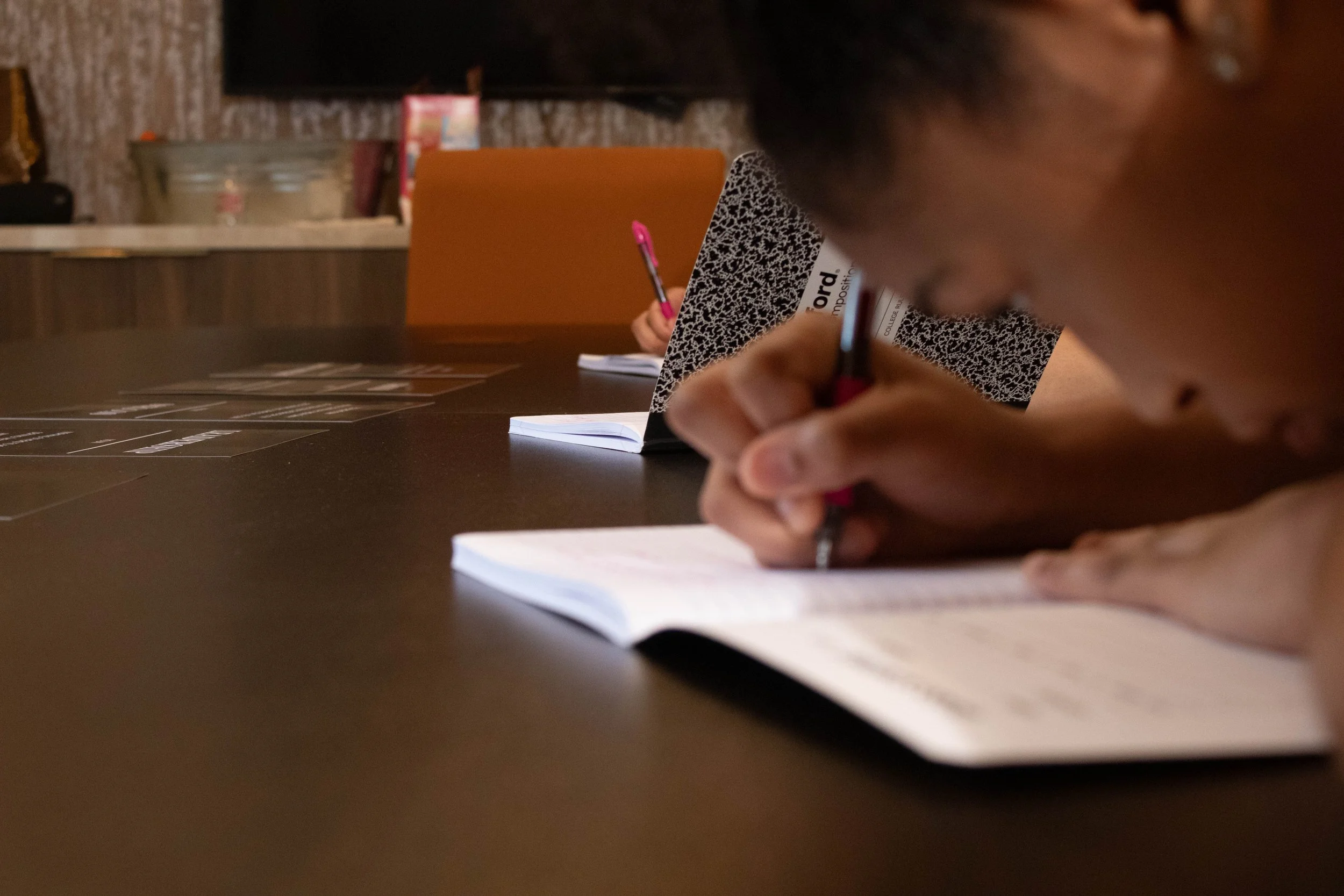 Person writing in a notebook in a dimly lit room, with another person in the background also writing, and objects like a menu and a tray visible on the table.