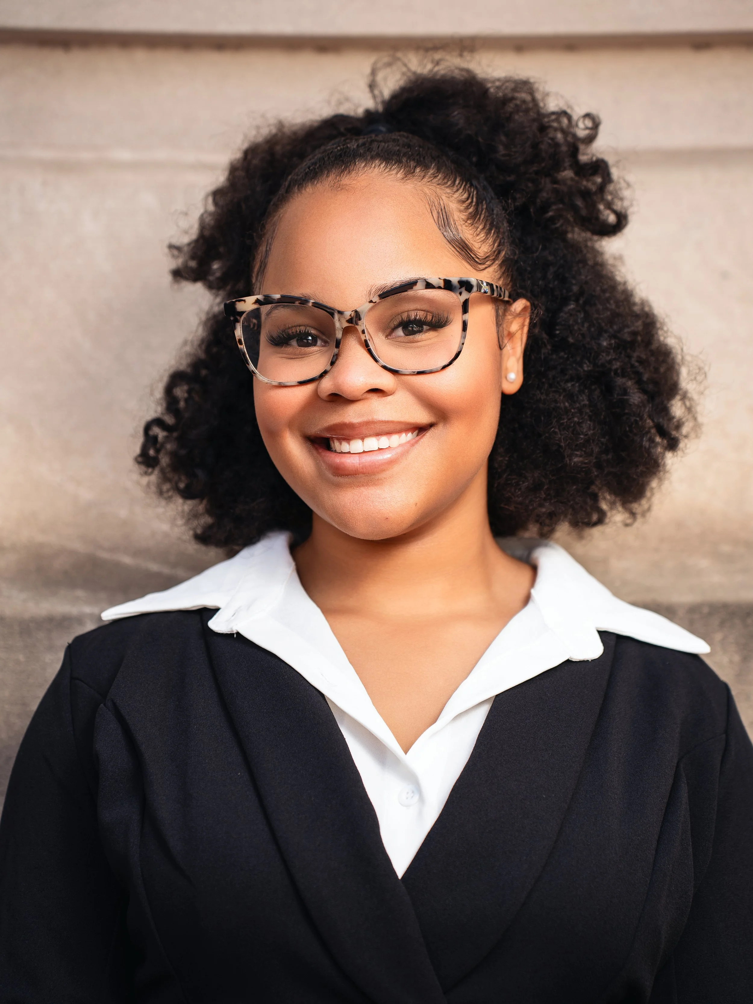 A young woman with curly black hair, glasses with a tortoise-shell pattern, wearing a white collared shirt and black blazer, smiling in front of a neutral background.