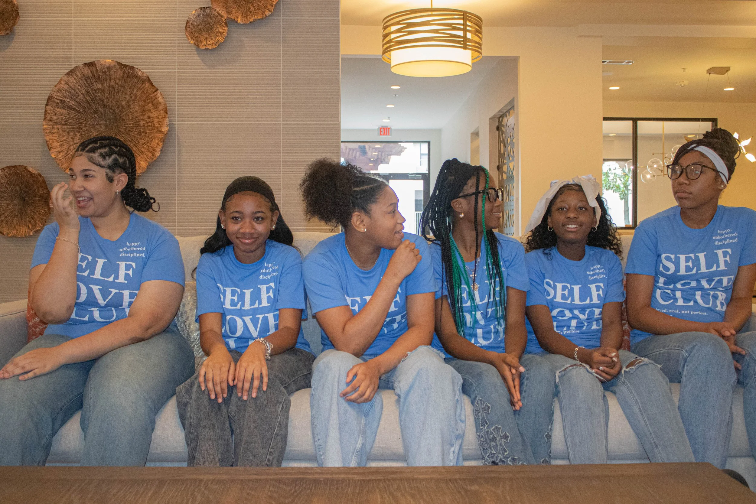 A group of six African American girls sitting on a couch, wearing matching blue T-shirts with the words "SELF LOVE CLUB" printed on them. They are in a brightly lit indoor space with modern decor, including a gray wall with large decorative wooden pi