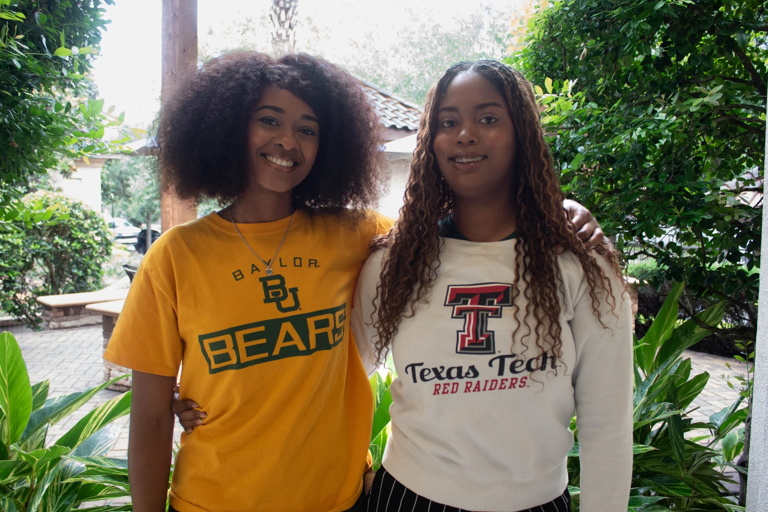 Two young women standing outdoors among green bushes, smiling at the camera. One is wearing a yellow Texas A&M shirt, and the other is wearing a white Texas Tech shirt.