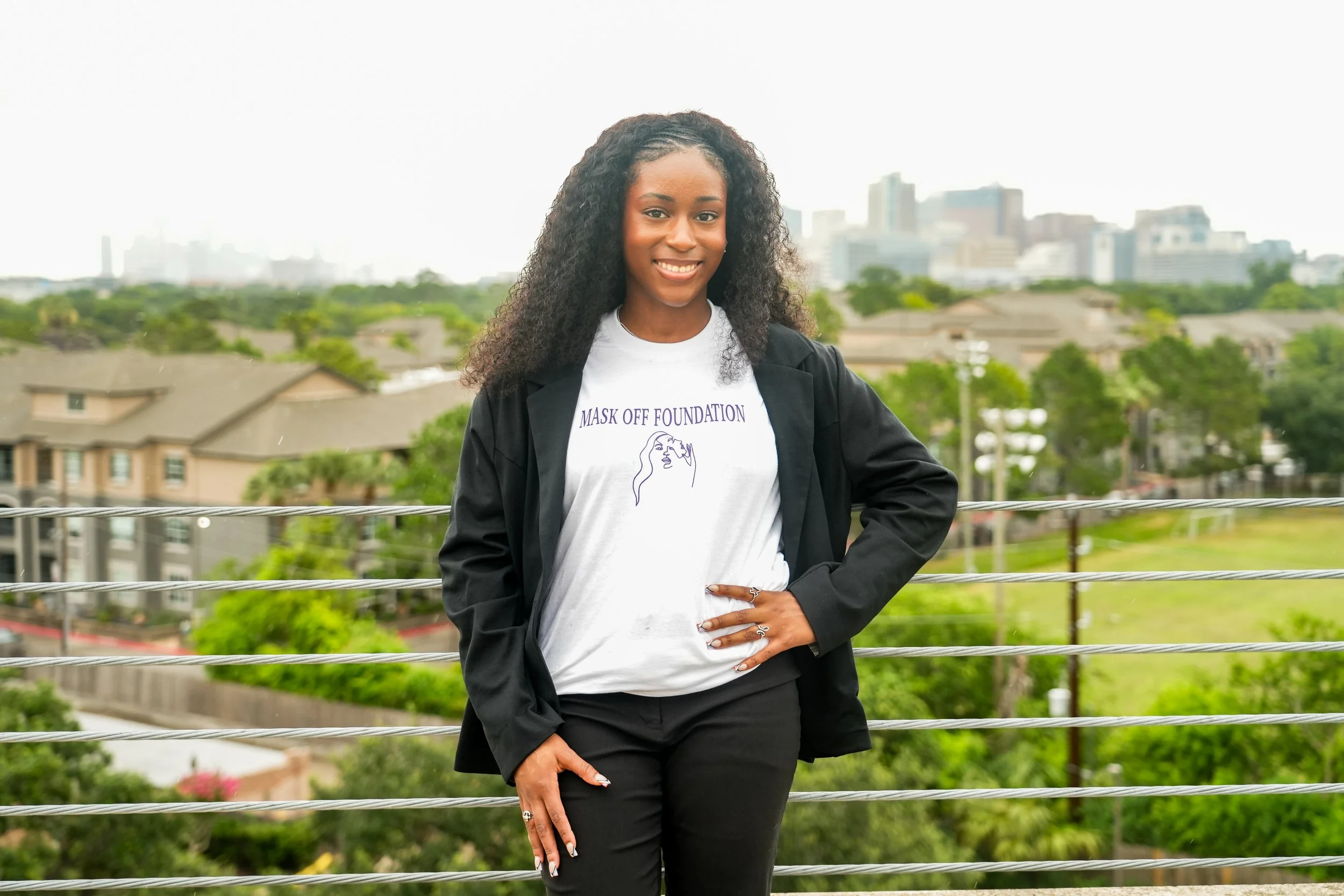A young woman with curly hair smiling and standing confidently outdoors on a balcony overlooking a cityscape with trees and buildings.