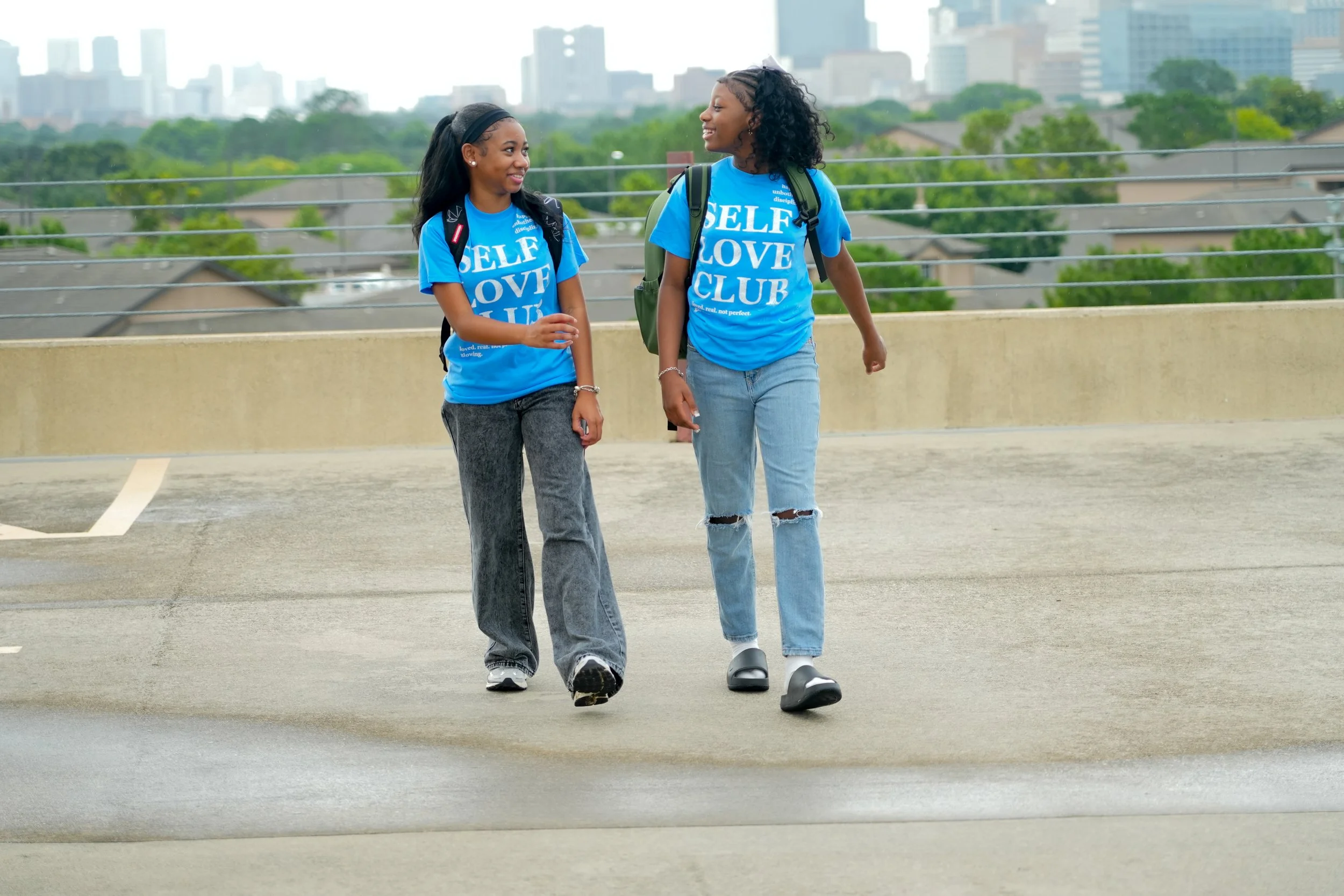 Two young women with backpacks walking and talking on a rooftop parking deck with a city skyline in the background.