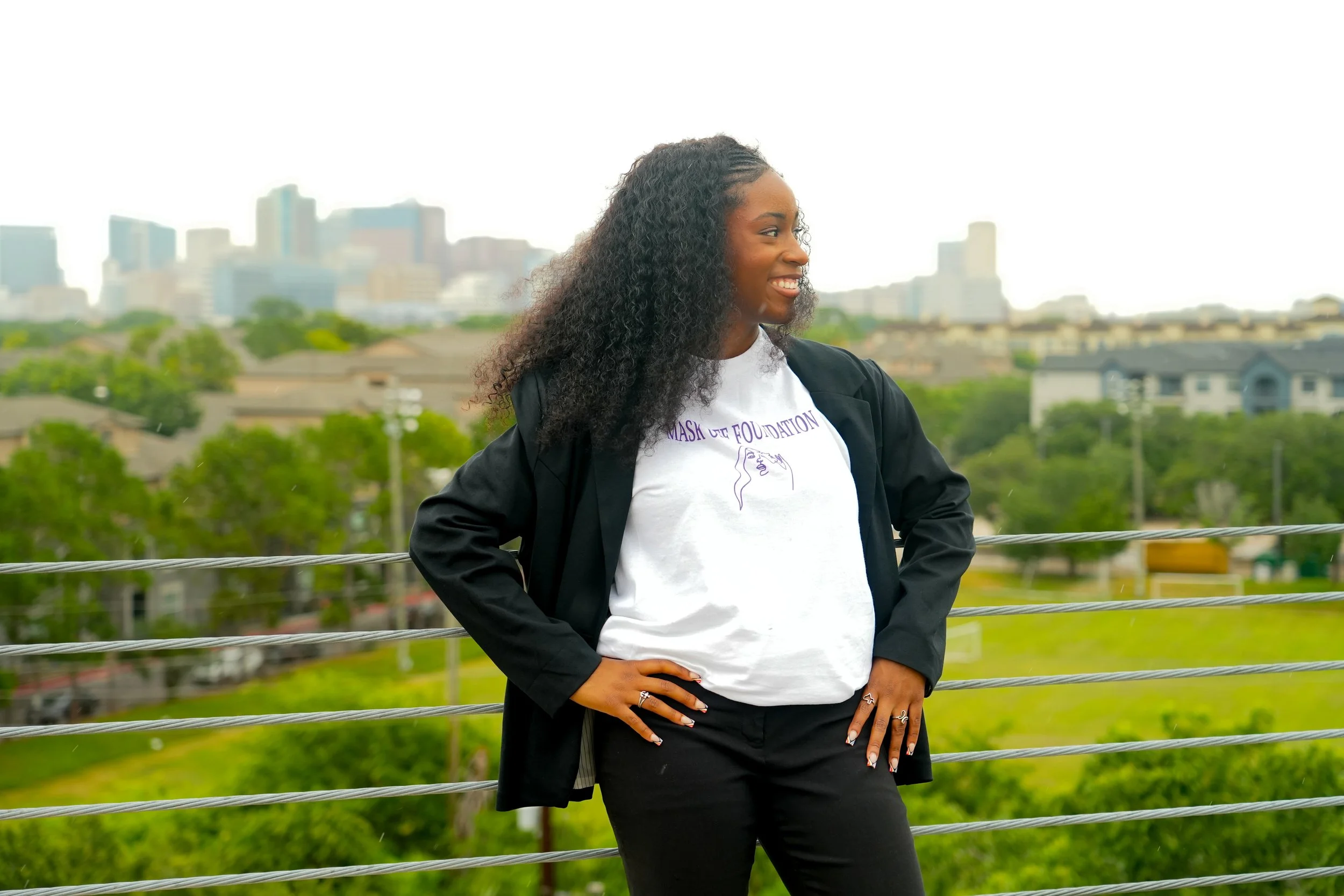 A woman with curly black hair smiling and standing outdoors with a city skyline in the background, wearing a white T-shirt and a black jacket.