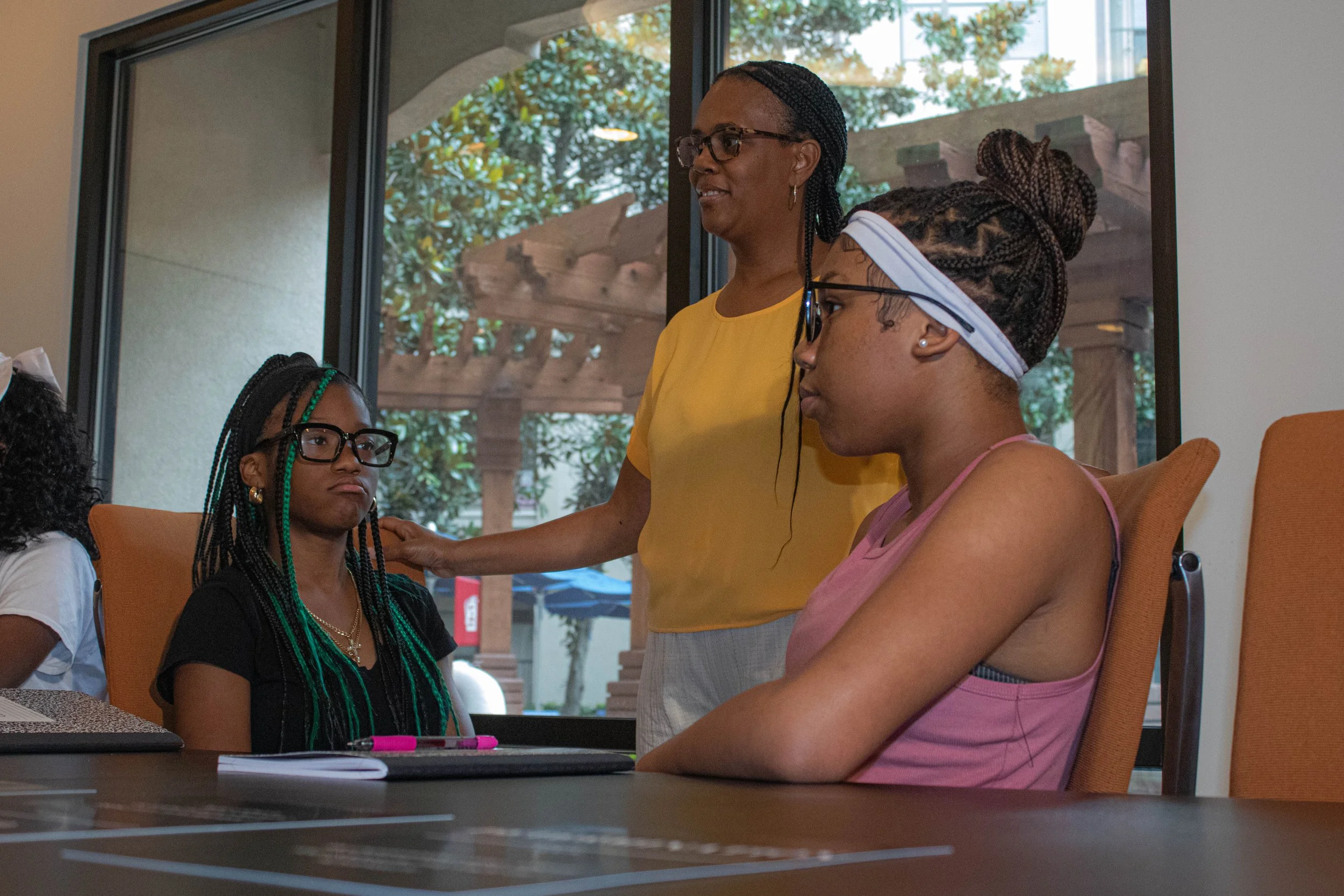 Four women, all wearing glasses, engaged in a discussion indoors with large windows showing trees outside. One woman in a yellow shirt appears to be speaking or listening intently, while the others listen and observe.