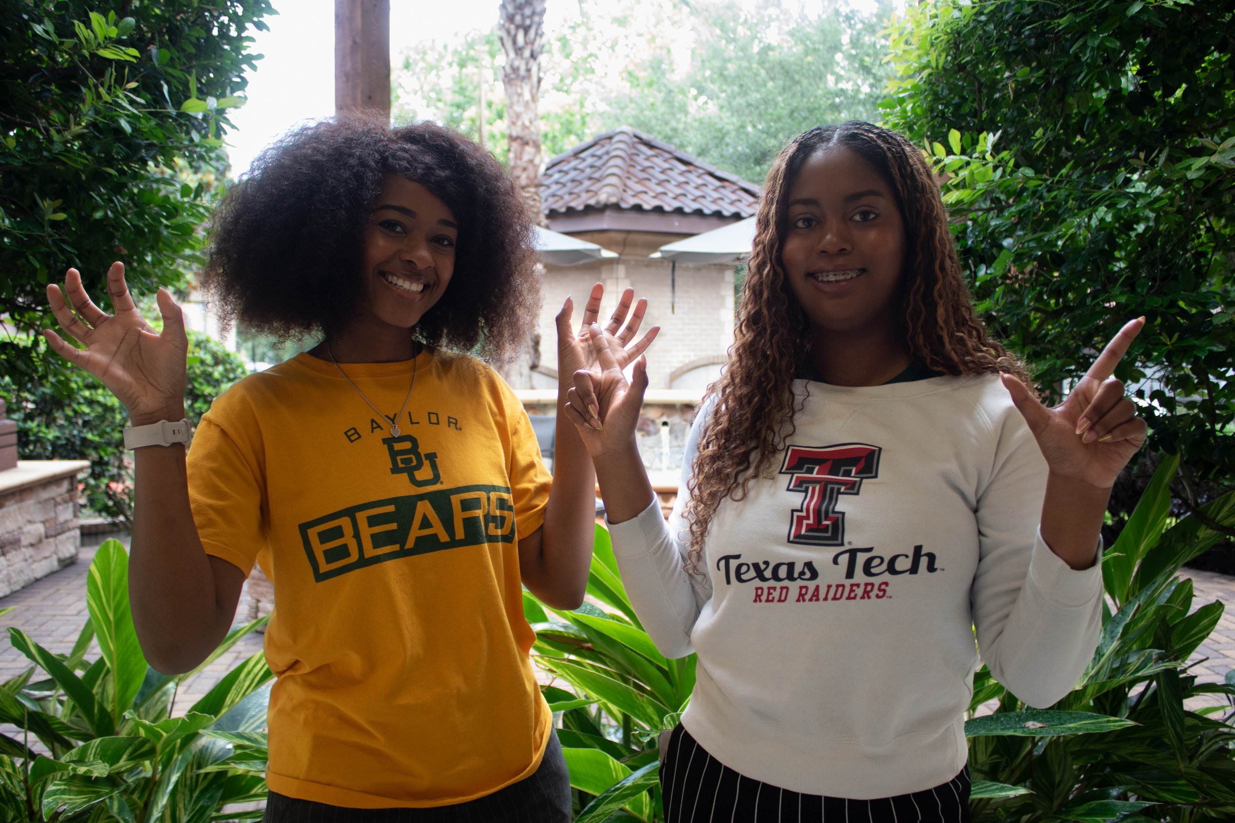 Two young women standing outdoors in a garden with lush green foliage, smiling, waving, and wearing college t-shirts.