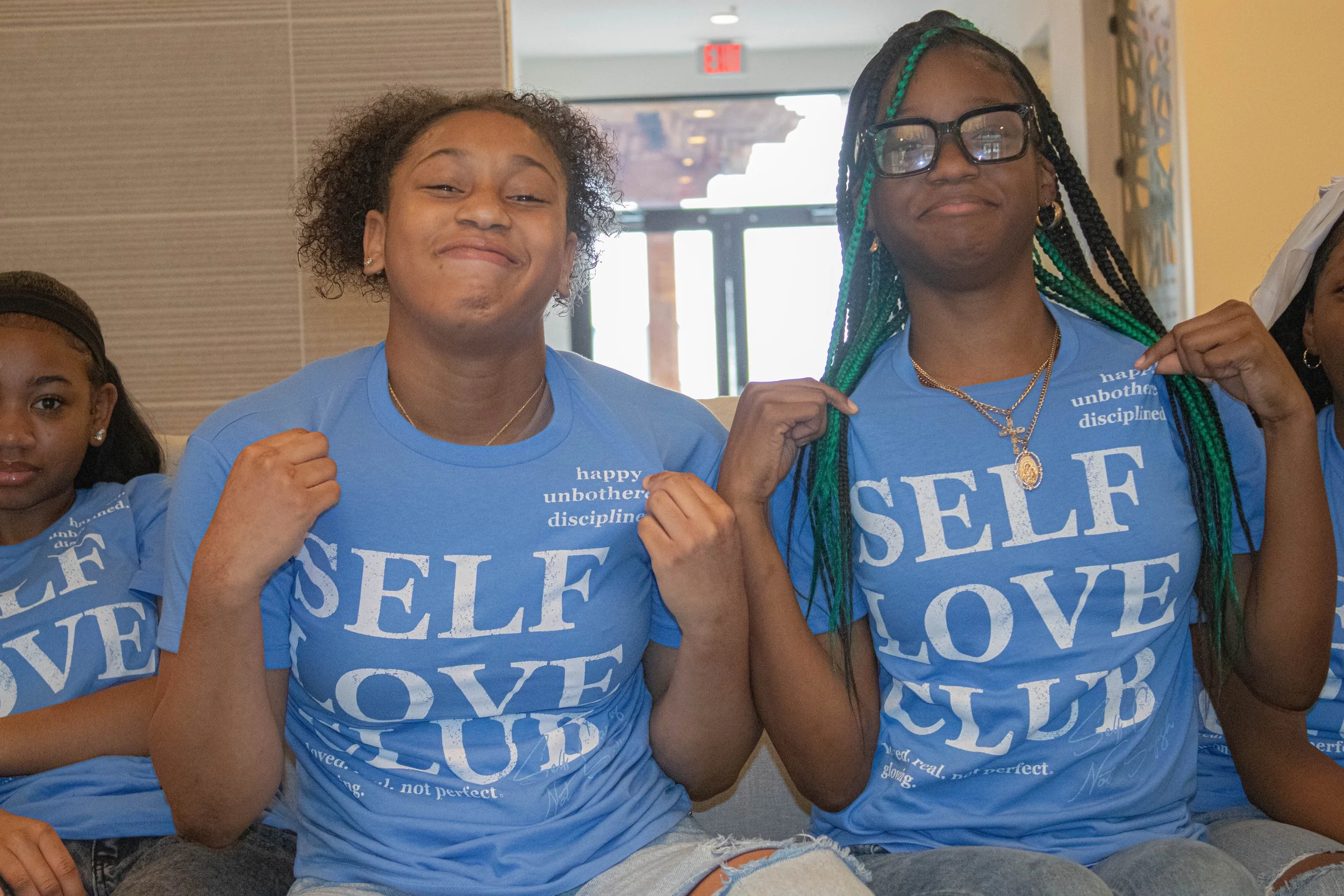 Group of young people sitting together indoors, wearing matching blue T-shirts with white text that says 'SELF LOVE' and other words. They are smiling confidently and making fists with their arms, showing strength and empowerment.