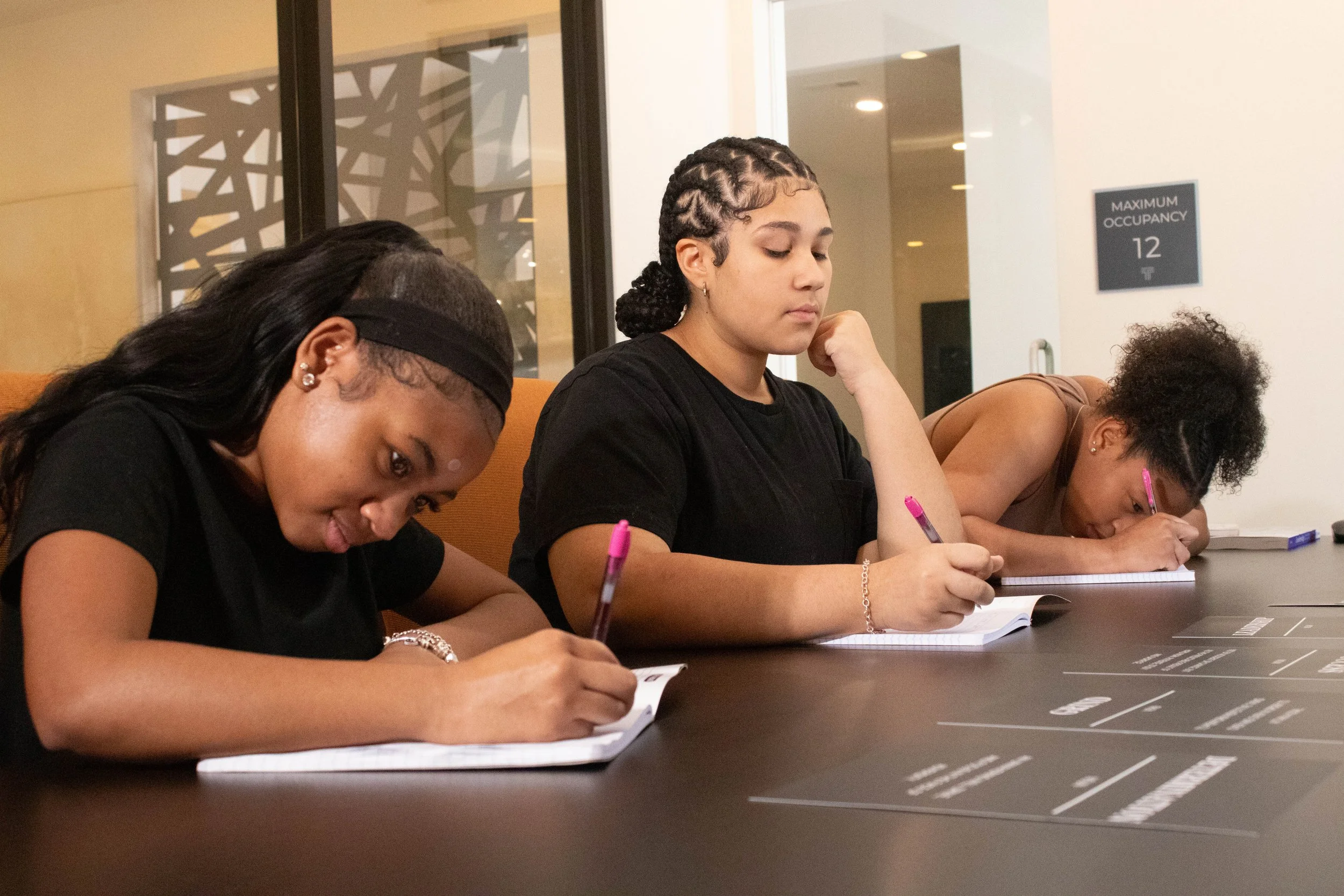 Four women sitting at a table, writing on notepads, with a sign in the background that says 'Maximum Occupancy 12'.