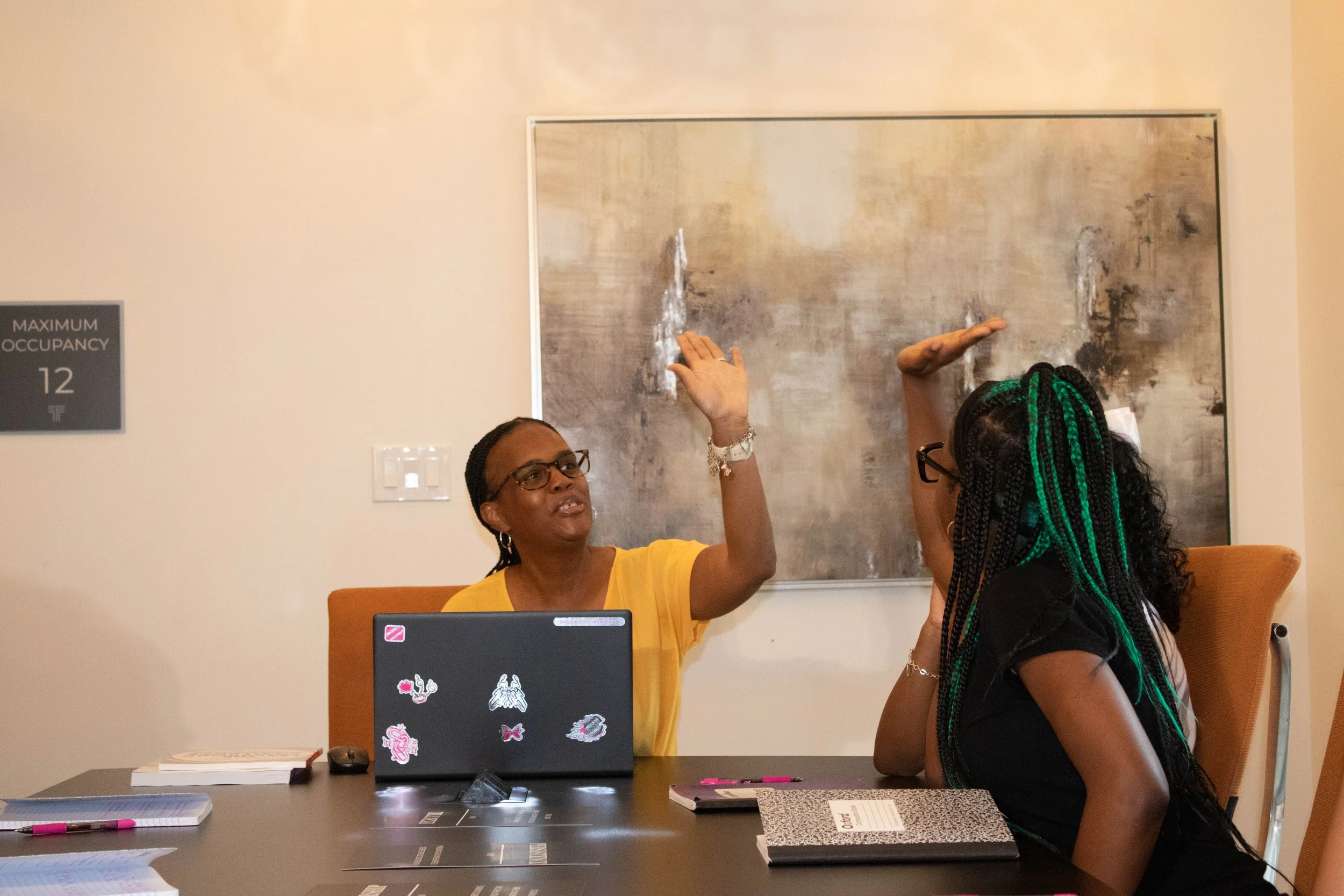 Two women sit at a conference table, engaging in a discussion. One woman with glasses and a yellow top is speaking and gesturing with her hands. The other woman with braided hair is listening attentively. The table has papers, notebooks, and sticky notes, and a laptop with colorful stickers. A large abstract painting is on the wall behind them, and a sign indicating maximum occupancy is visible.