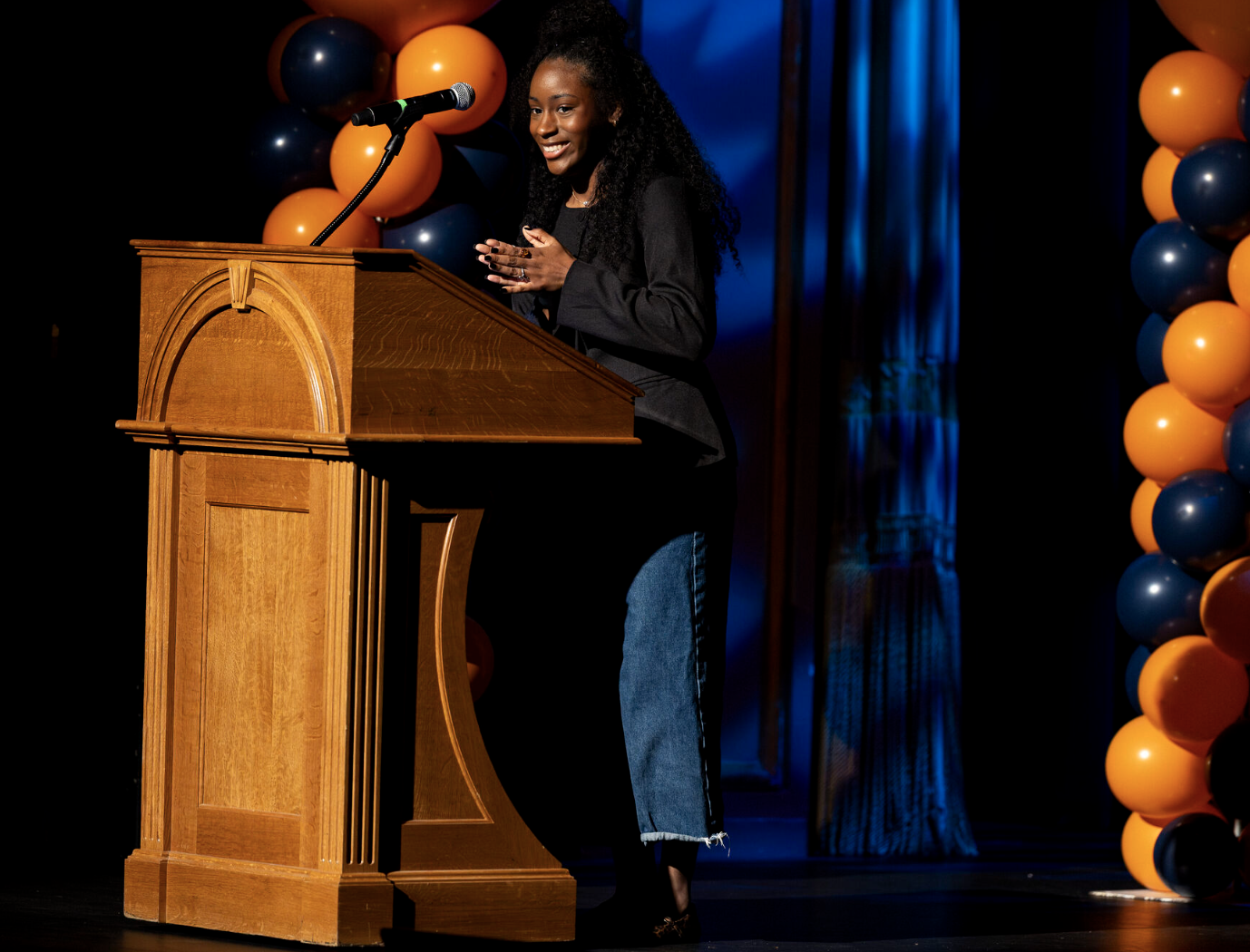 A young woman standing at a wooden podium on stage, smiling, with balloons in orange, black, and blue behind her, during an event or speech.