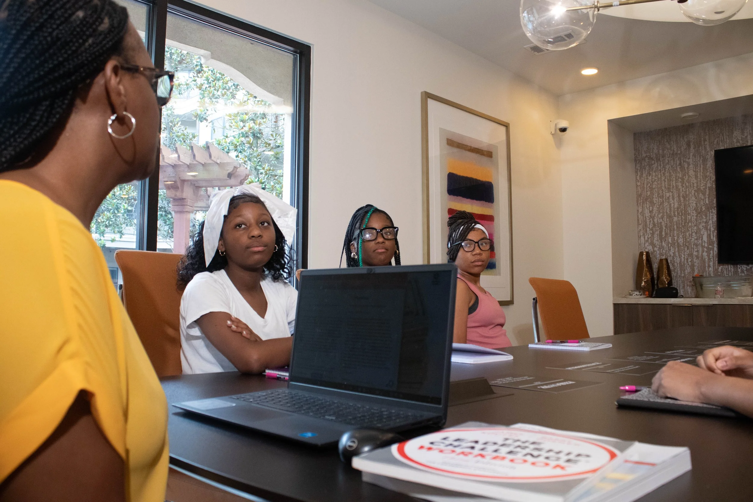 Four women seated around a conference table in a meeting room, with a woman in a yellow top speaking or listening, and three younger women with glasses, one with a white head covering, all paying attention, laptops and notebooks on the table, large w